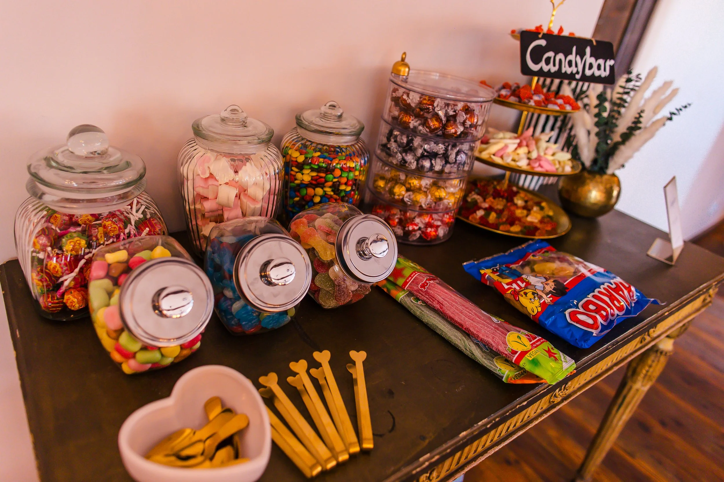 Verschiedene Süßigkeiten in Gläsern und Schalen auf einem Tisch, inklusive Gummibärchen, Zuckerstangen, Fruchtgummi und Bonbons, mit einer Candybar-Beschilderung im Hintergrund.