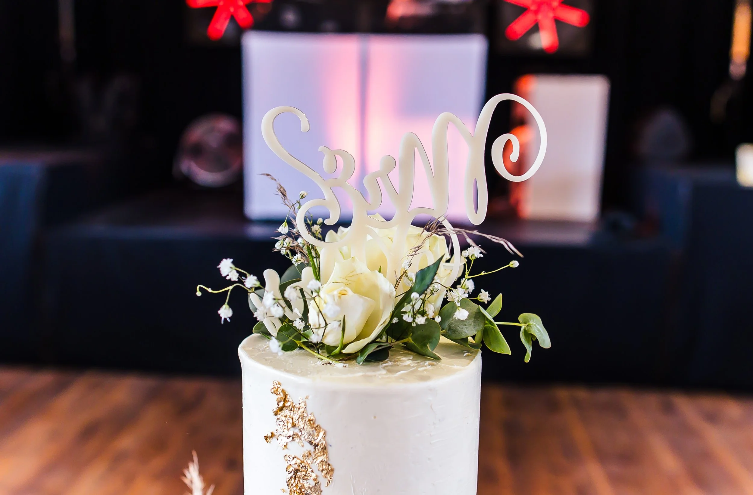 Weiße Hochzeitstorte mit Blumen und einem silbernen Schriftzug, der 'Sasno' sagt, auf der Oberseite. Im Hintergrund ein Geschenk mit rotem Schleifenbändchen.
