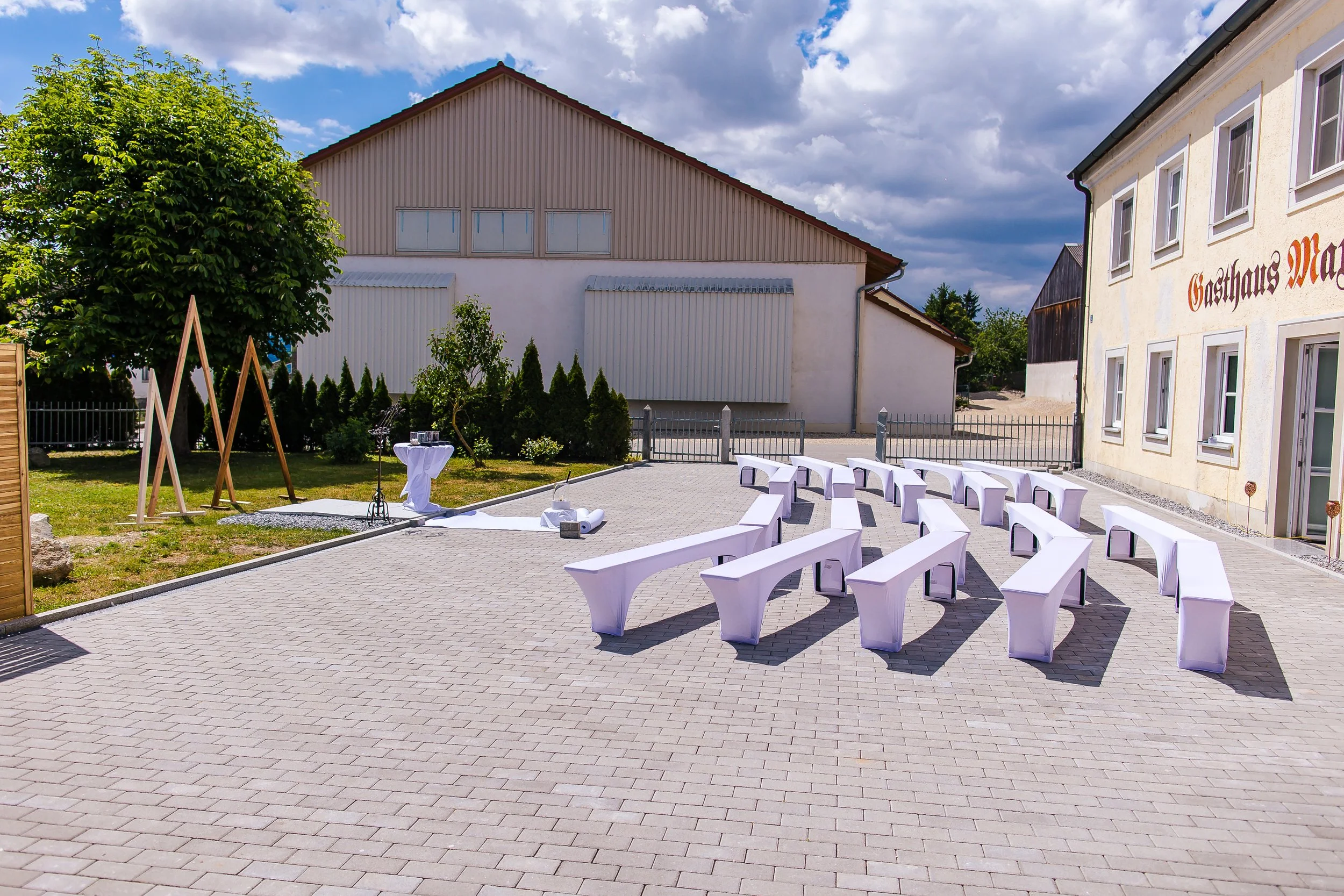 Außerhalb eines Gebäudes sind weiße, mit Tüchern bedeckte Stühle und Bänke für eine Zeremonie auf einem gepflasterten Platz arrangiert. Es gibt eine kleine Bar mit Getränken auf einem hohen Tisch und eine Klavierrequisite in der Nähe. Im Hintergrund 