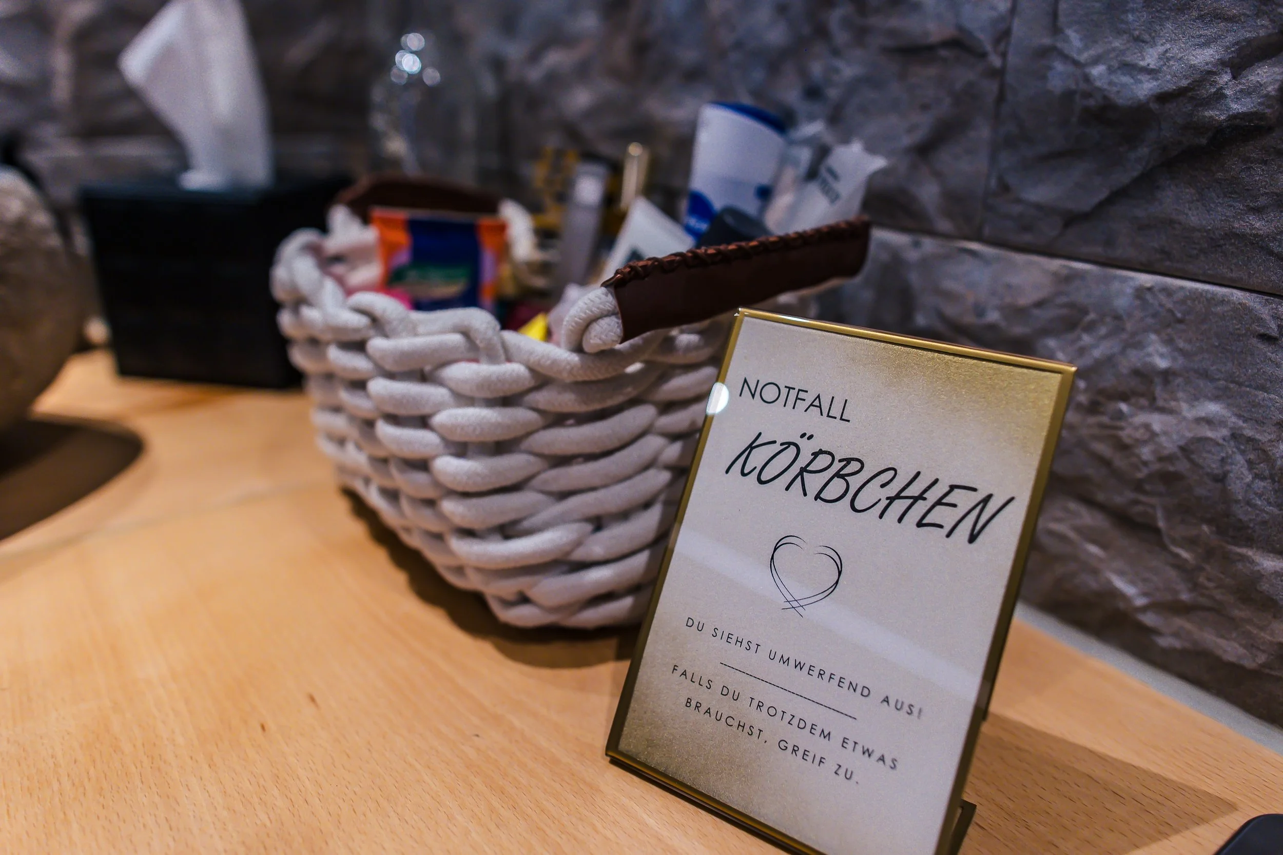 Eine Kiste mit verschiedenen Gegenständen und ein Schilder mit der Aufschrift 'Notfall Körbchen' auf einem Holztisch in einer Umgebung mit Steinwand.
