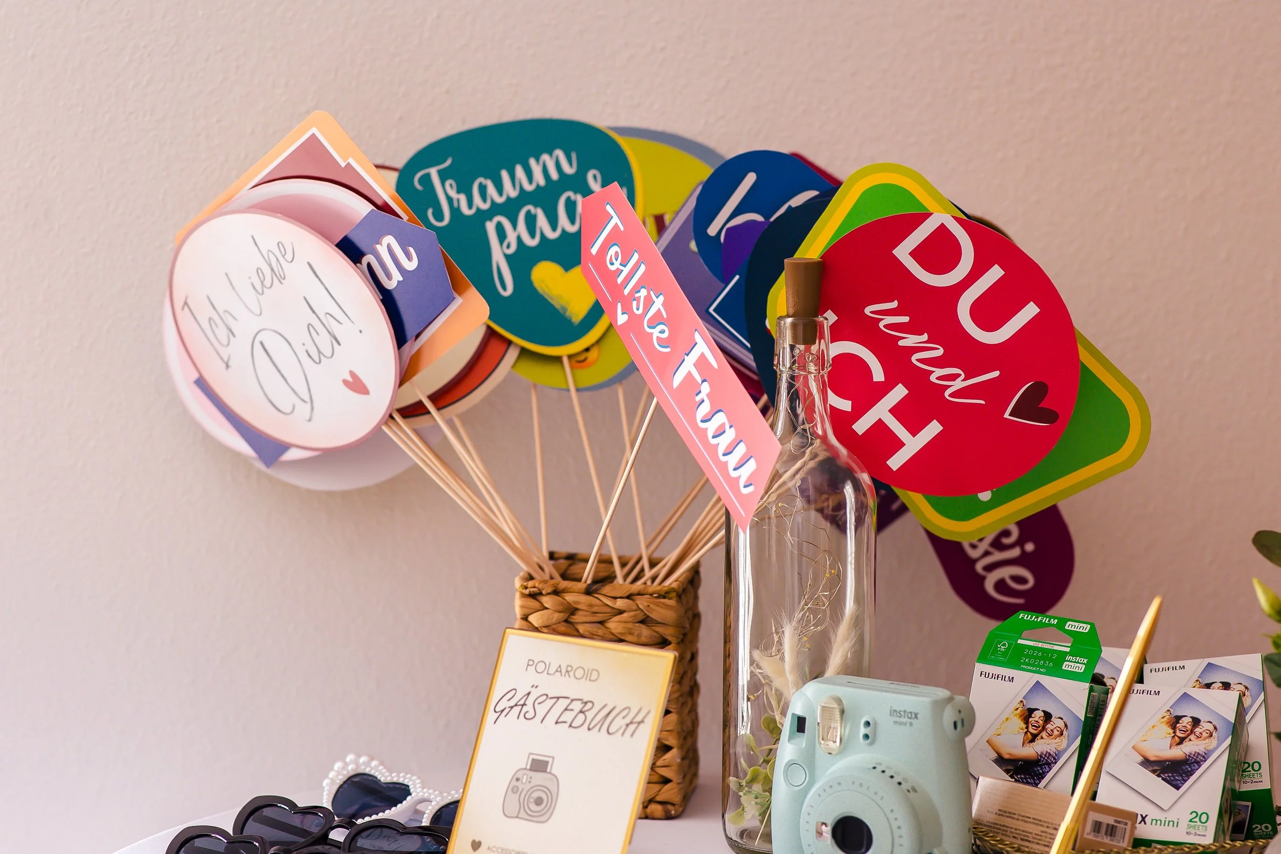 Bunte Fotoschilder mit Liebesbotschaften wie 'Ich liebe Dich', 'Traum Pace', 'Du und Ich' auf Holzstäbchen in einem Korb, eine Polaroid Gäste Buch, eine Instax Kamera, Sonnenbrillen und Fotofilme auf einem Tisch.