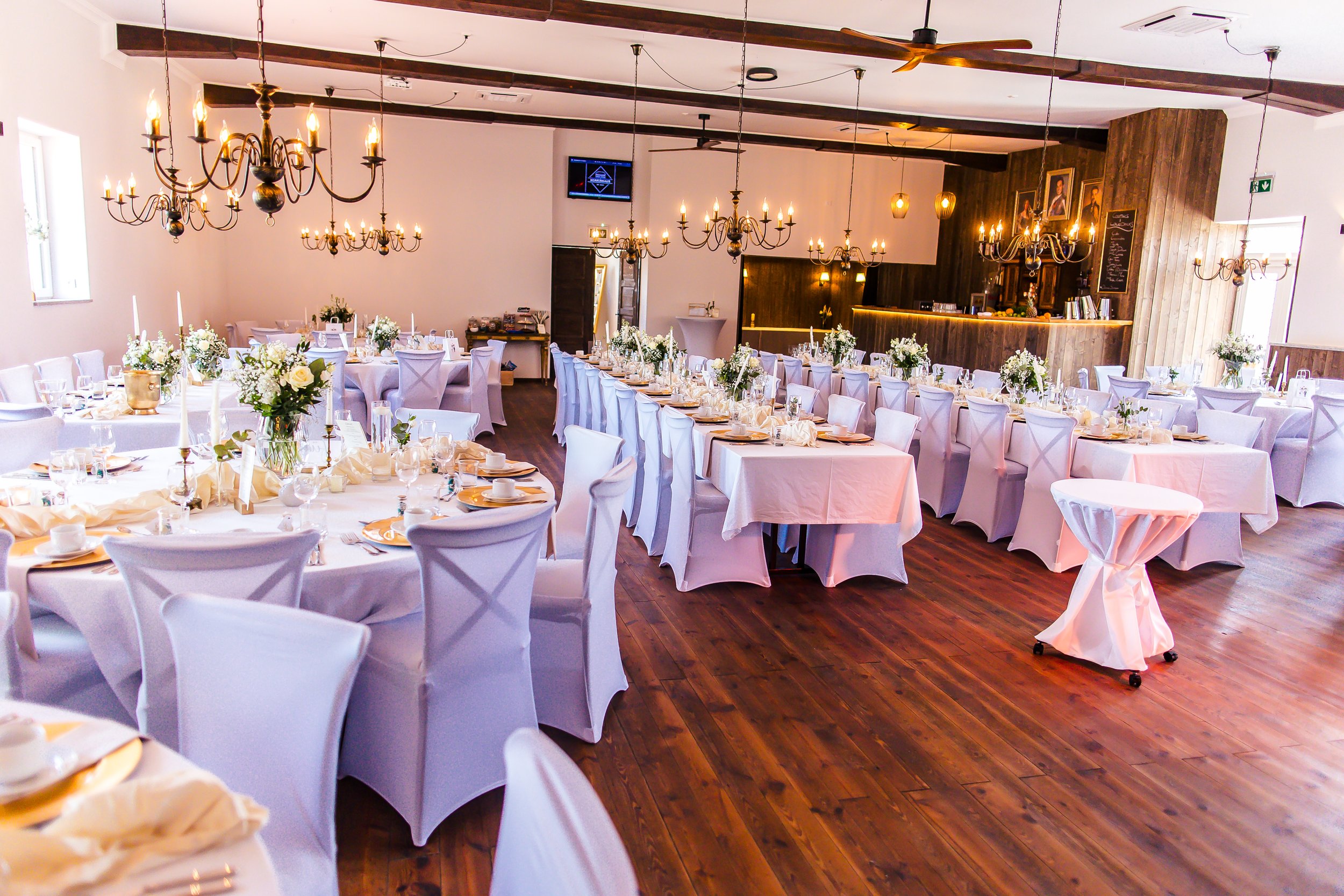 Elegantes Festsaal mit lange Tische mit weißen Tischdecken, Blumenarrangements, Kerzen und festlicher Besteckdekoration, dekoriert für eine Hochzeit oder Feier, Vielzahl von Stühlen mit weißen Bezügen, Holzfußboden, Kronleuchter an der Decke, bar mit