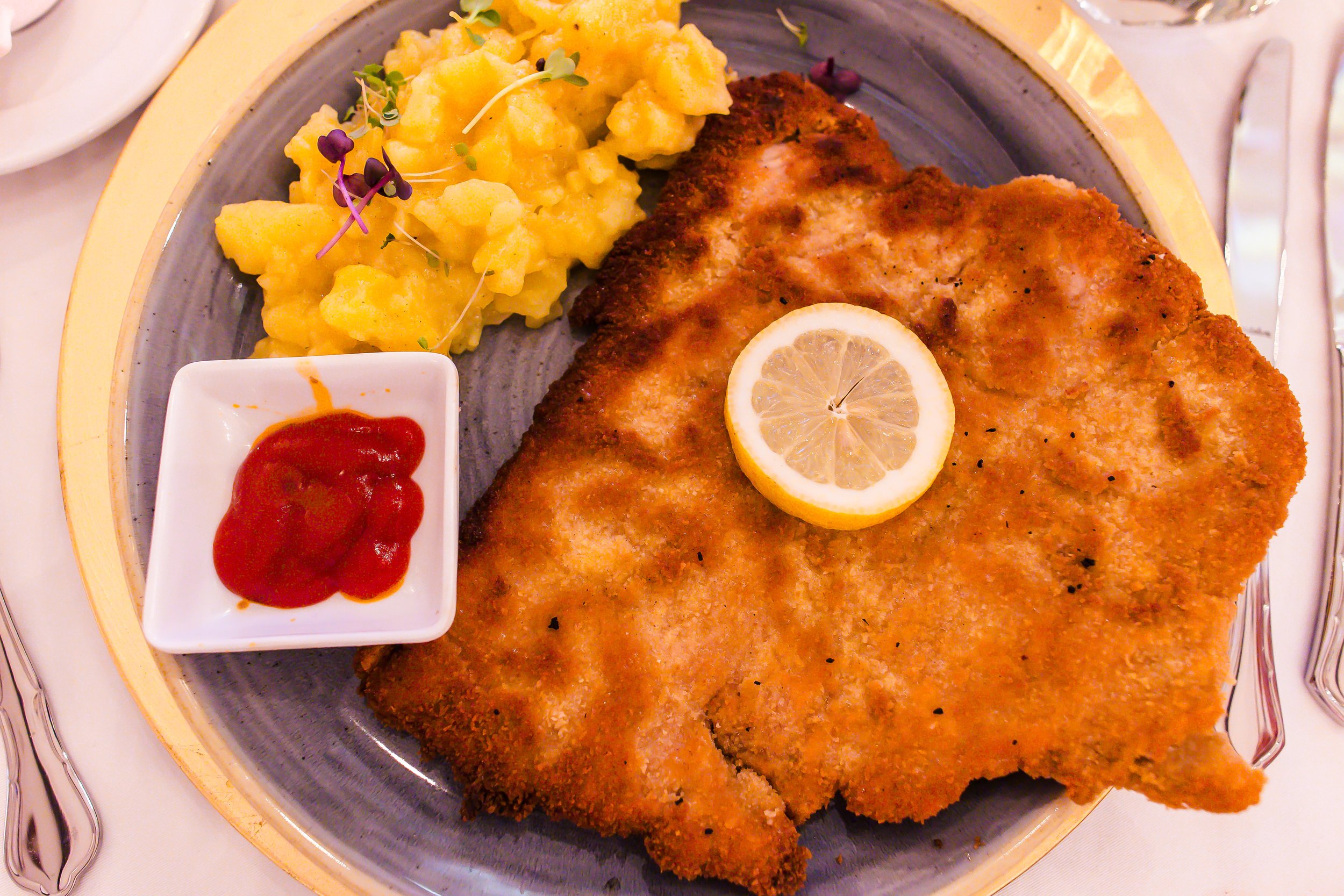 Schnitzel mit Zitronenscheibe, Kartoffelsalat mit Microgreens, Ketchup auf weißem Teller, auf einem grauen und goldenen Teller serviert.