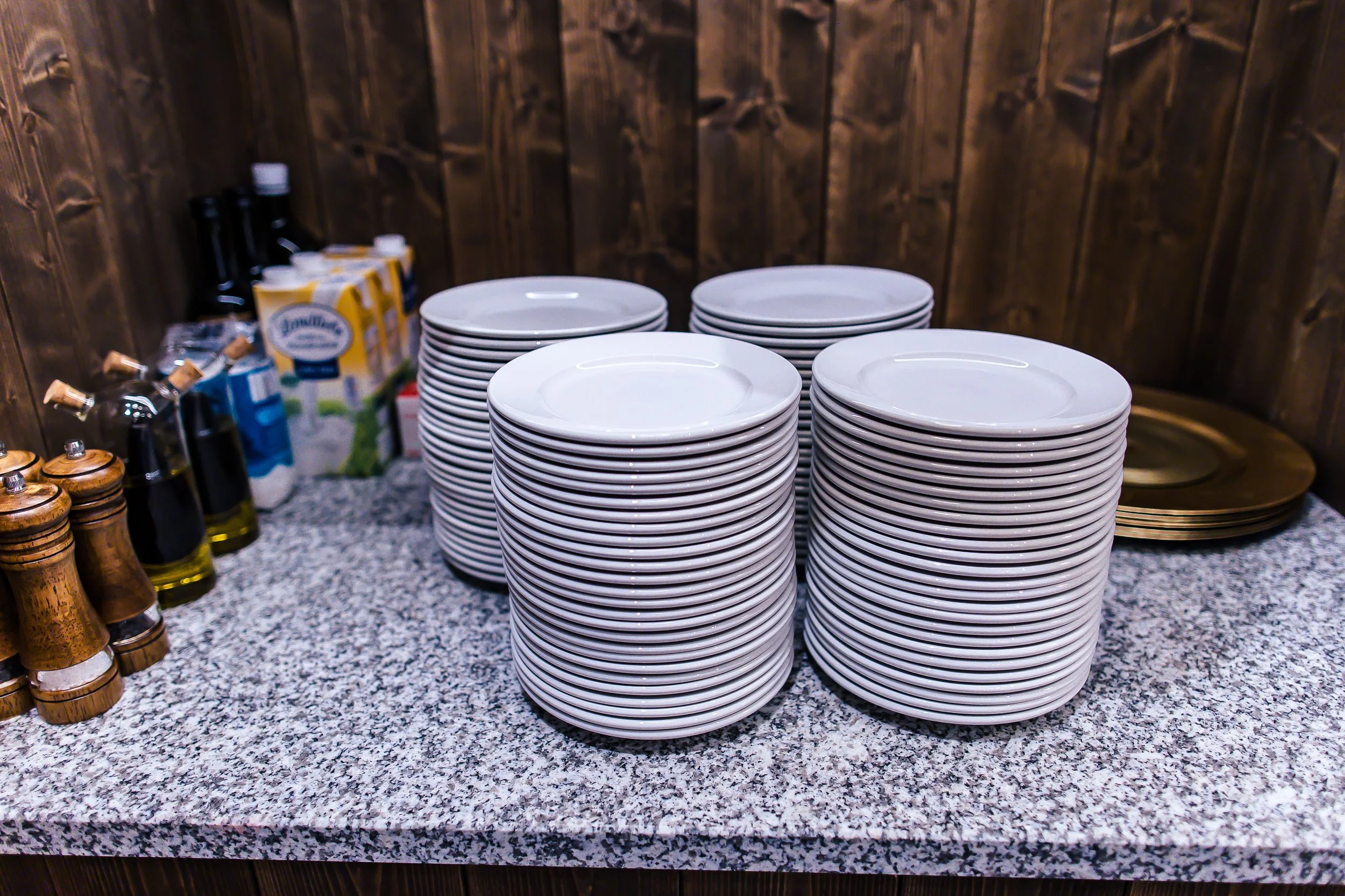 Ein Stapel weißer Teller auf einer Speisetafel, im Hintergrund sind Gewürzstreuer, Ölflaschen und Käsesorten sichtbar.