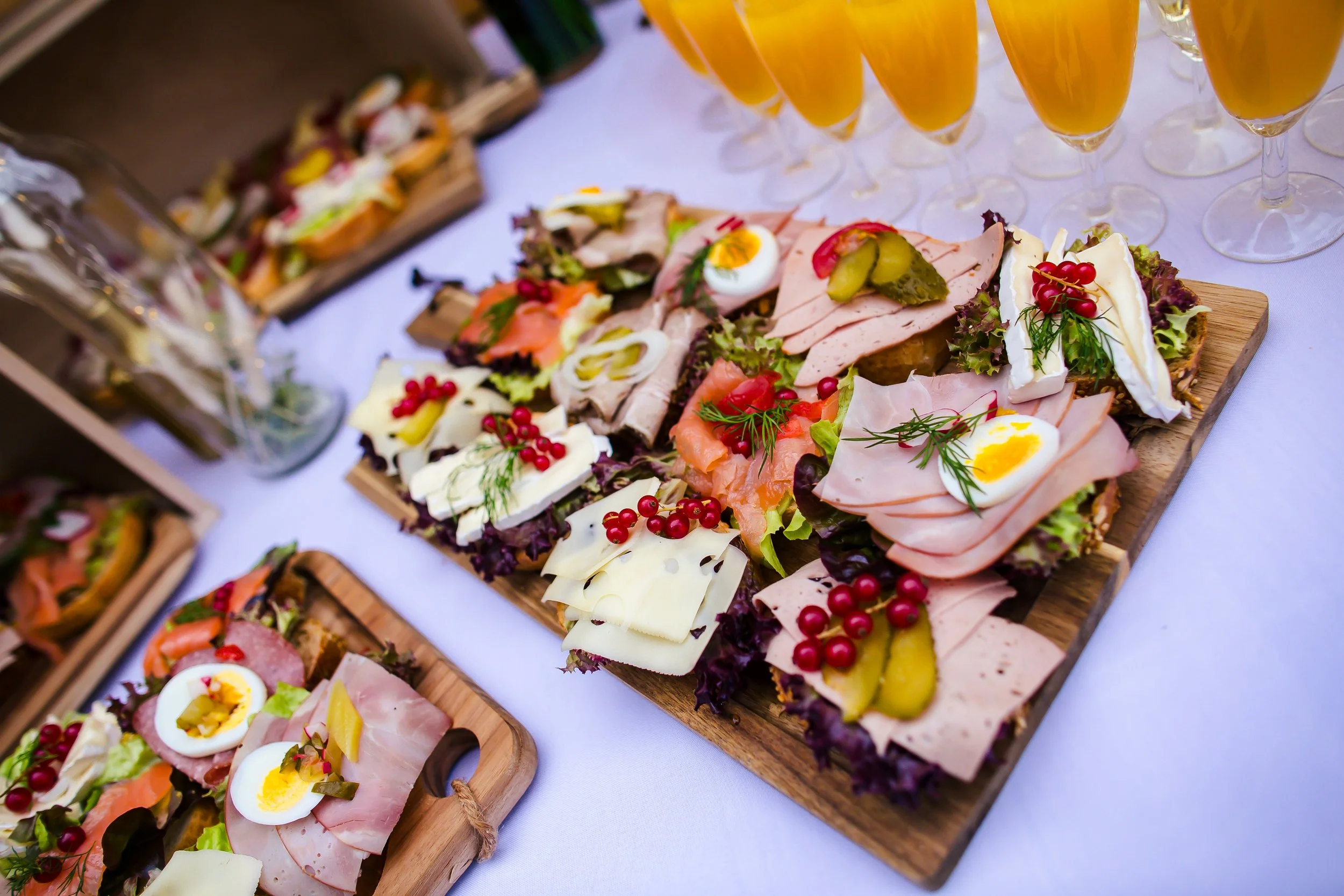 Verschiedene belegte Brote mit Ei, Aufschnitt, Käse, Gurken, Kirschen und Salat auf Holzplatten, daneben Sektgläser.