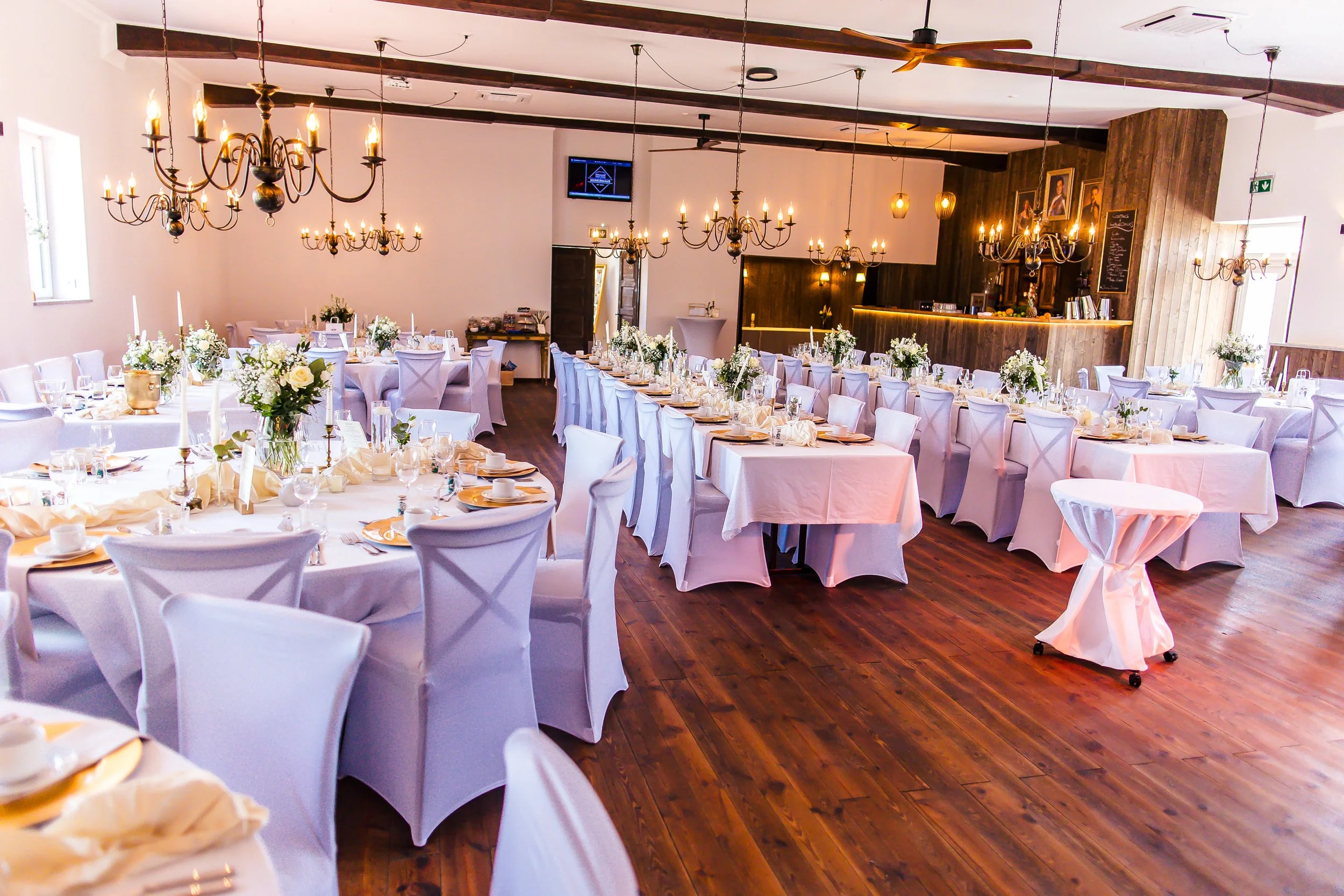 Eleganter festlich gedeckter Saal mit weißen Tischdecken, kristallenen Gläsern, weißen Stuhlhussen, mittelgroßen Blumenarrangements und antiken Kronleuchtern, vorbereitet für eine Hochzeit oder Feier.