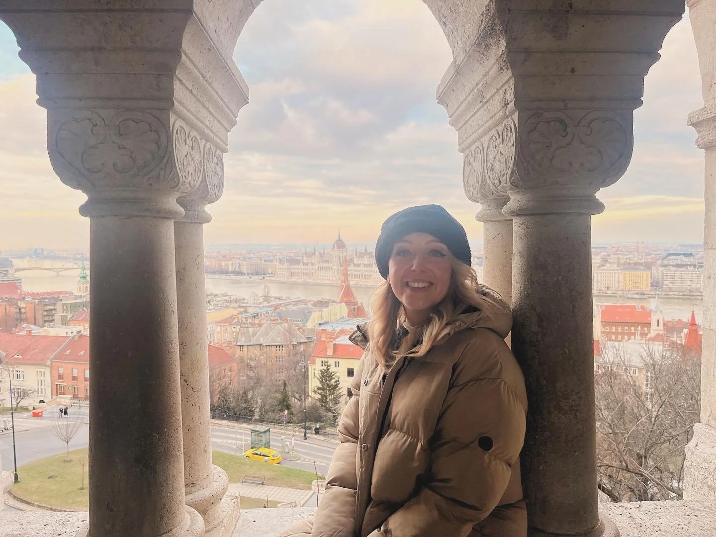 A woman smiling wearing a brown puffer jacket and black beret, sitting between stone columns with a cityscape view of Budapest, Hungary in the background, including the Danube River and Parliament building.
