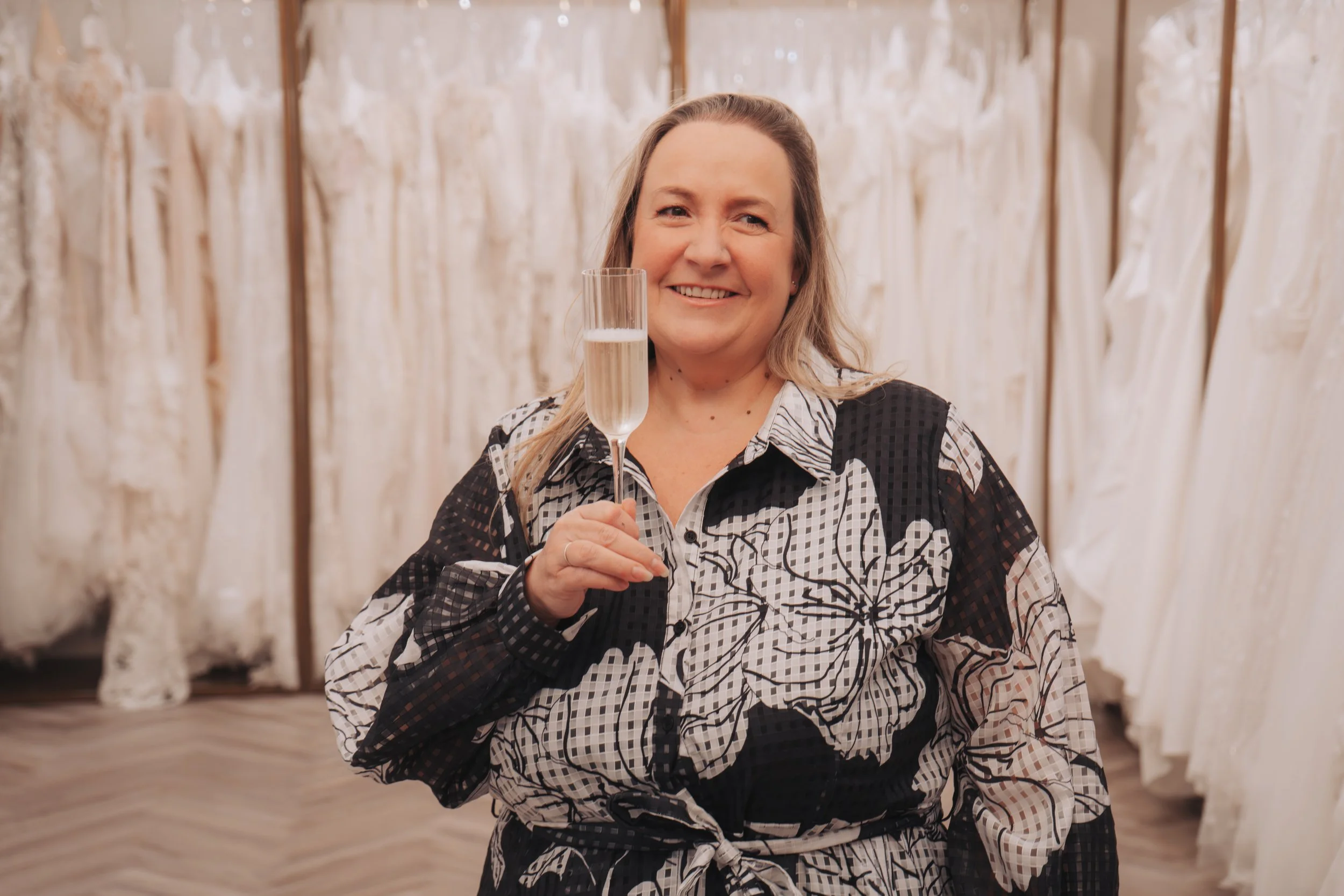 Owner of the bridal store smiling with a glass of bubbly in hand stood in front of the beautiful bridal gowns