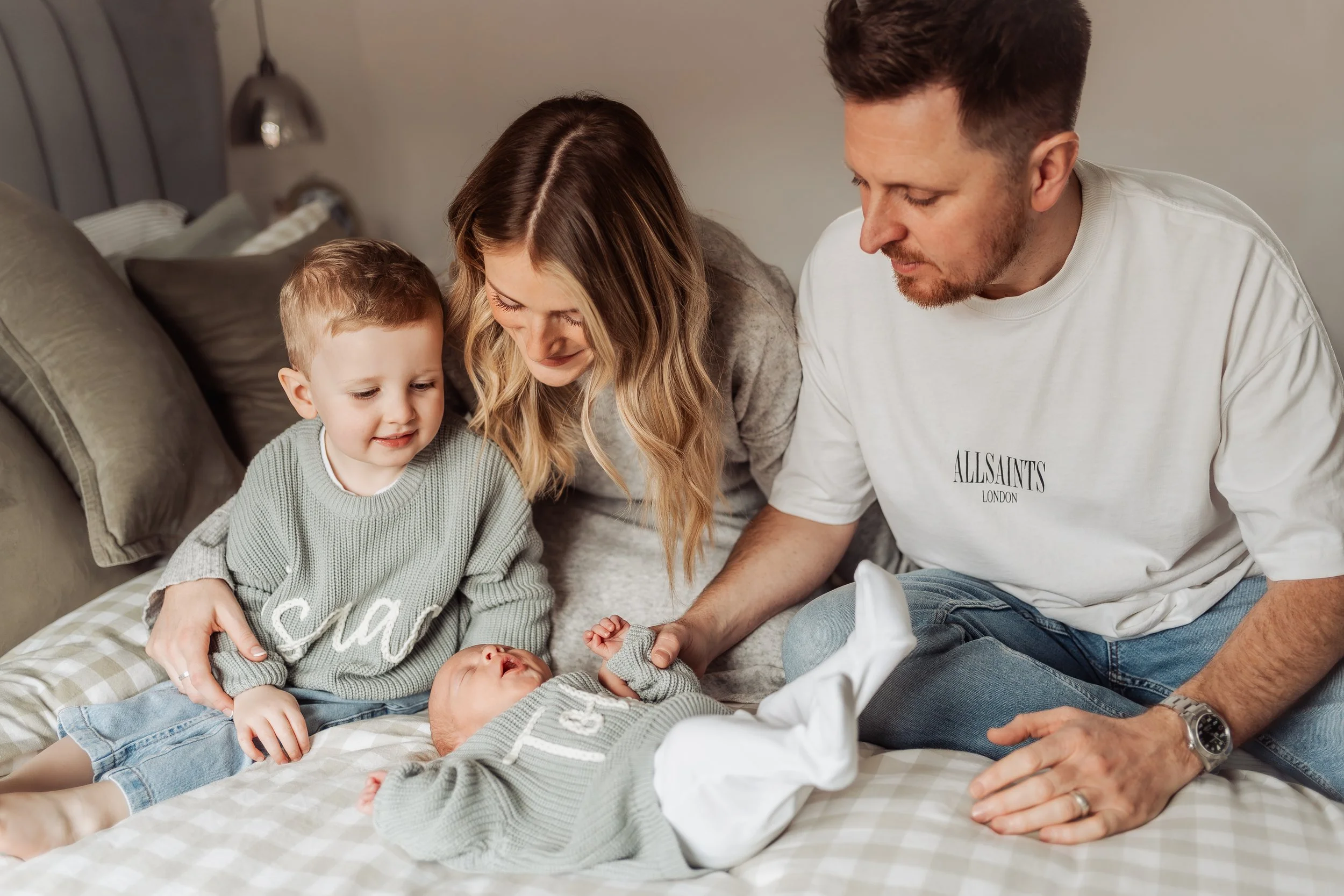 A family gathering around a newborn baby on a bed, with a young boy, mother, father, and a newborn, in a cozy bedroom setting.