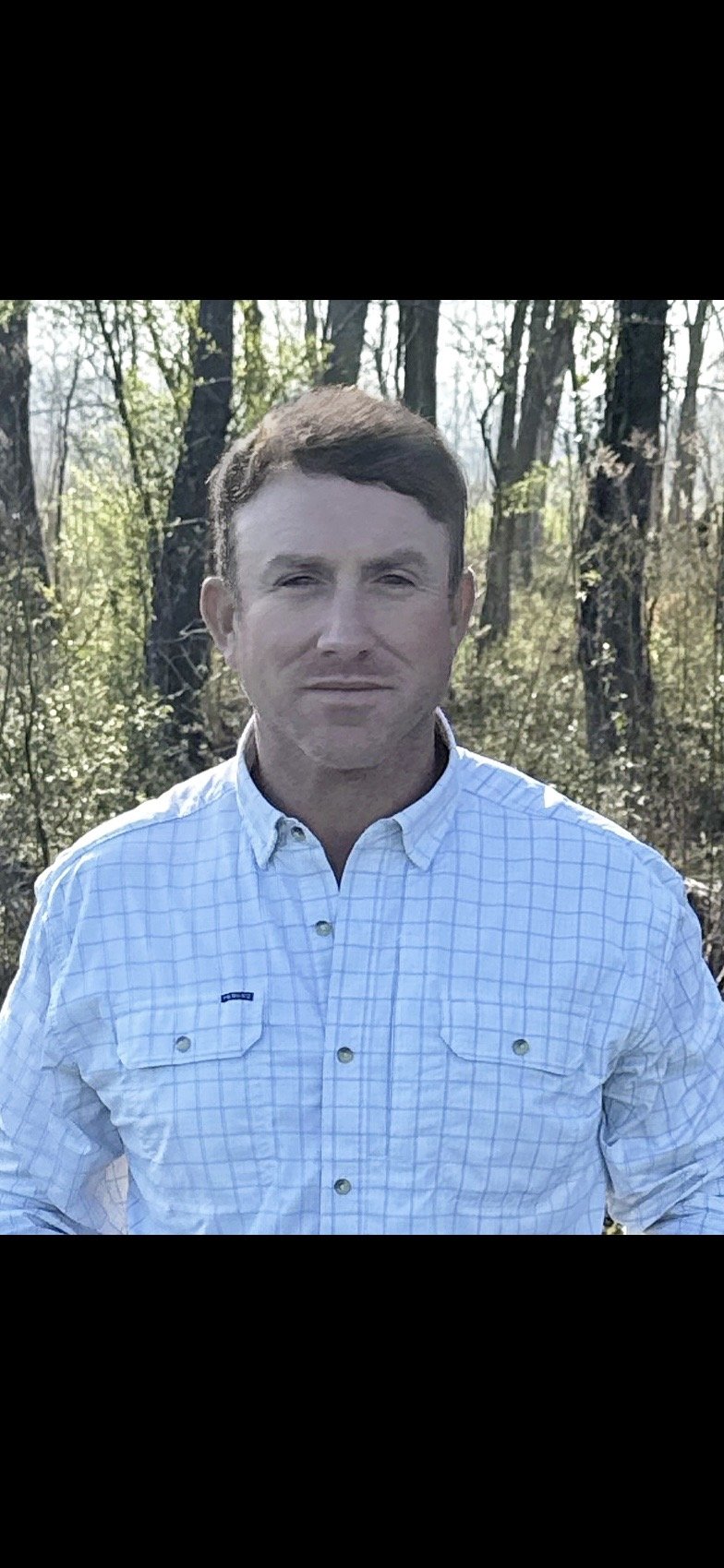 A man with short brown hair wearing a light blue plaid button-up shirt standing outdoors in a wooded area with trees in the background.