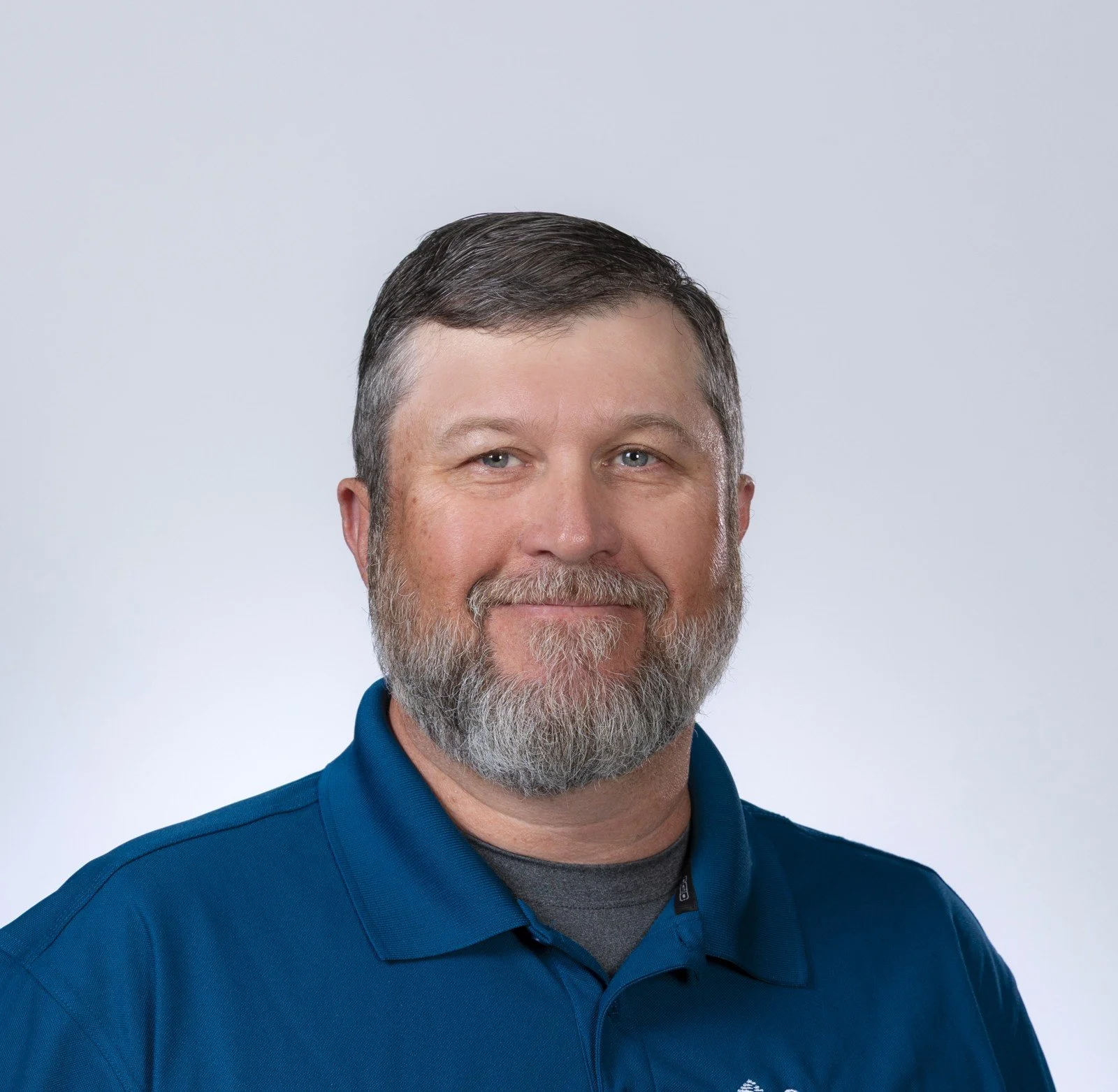 Headshot of a middle-aged man with gray hair, beard, and blue eyes, wearing a blue collared shirt, against a plain light gray background.