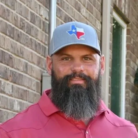 A man with a gray beard wearing a gray baseball cap with a Texas-themed logo and a maroon polo shirt, standing outdoors near a brick building.