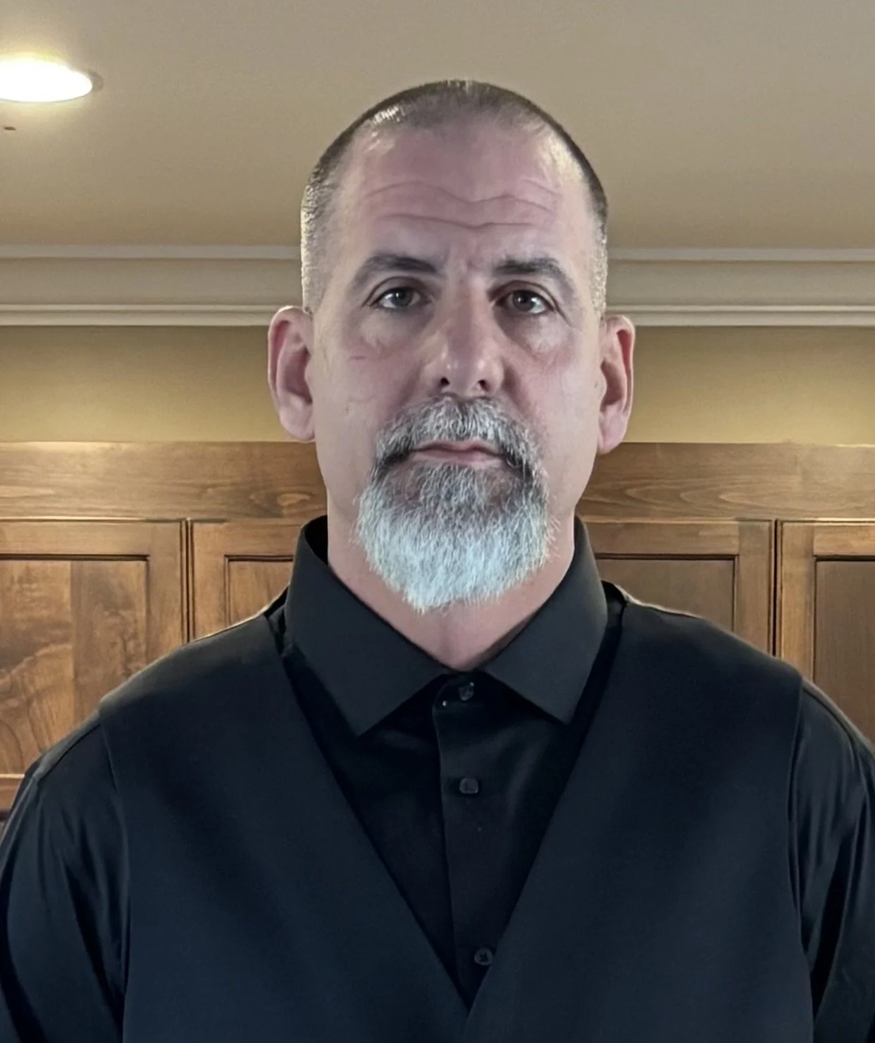 A middle-aged man with a bald head, gray beard, and mustache wearing a black collared shirt in front of wooden cabinets.