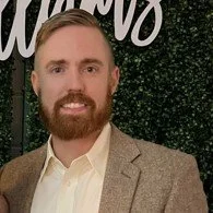 A man with a beard and light brown hair, wearing a beige blazer and white shirt, standing in front of a green foliage backdrop.