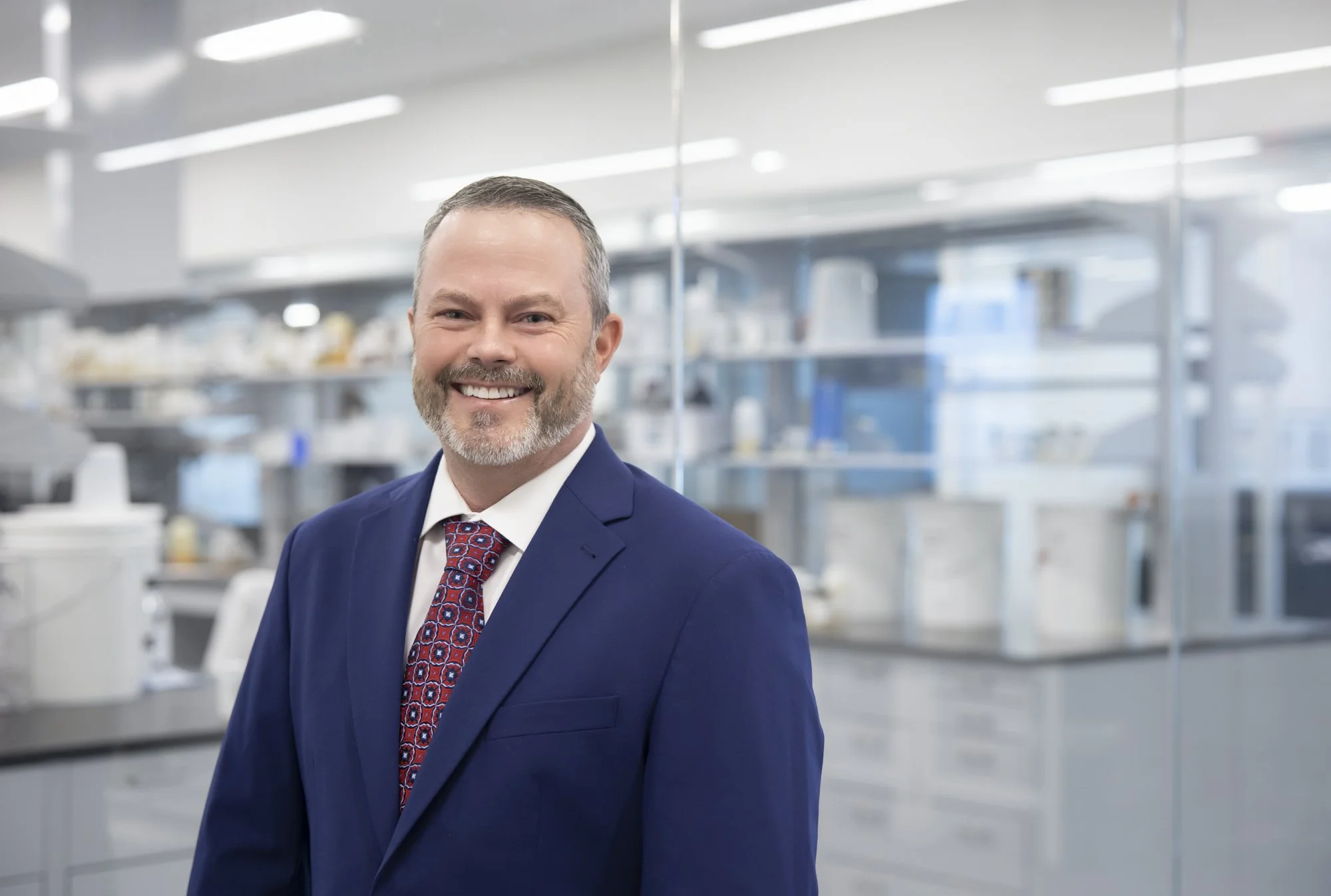 A smiling man in a blue suit and patterned tie standing in a bright, modern laboratory with shelves of scientific equipment in the background.