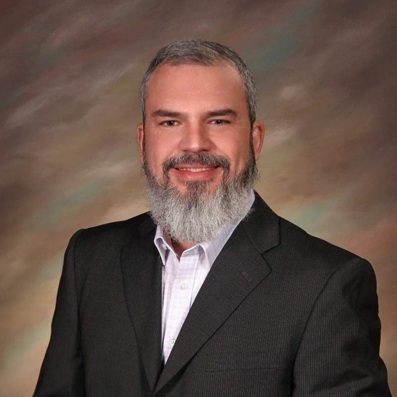 A man with a beard and short hair wearing a black suit and white shirt, smiling, with a professional studio background in shades of brown and gray.
