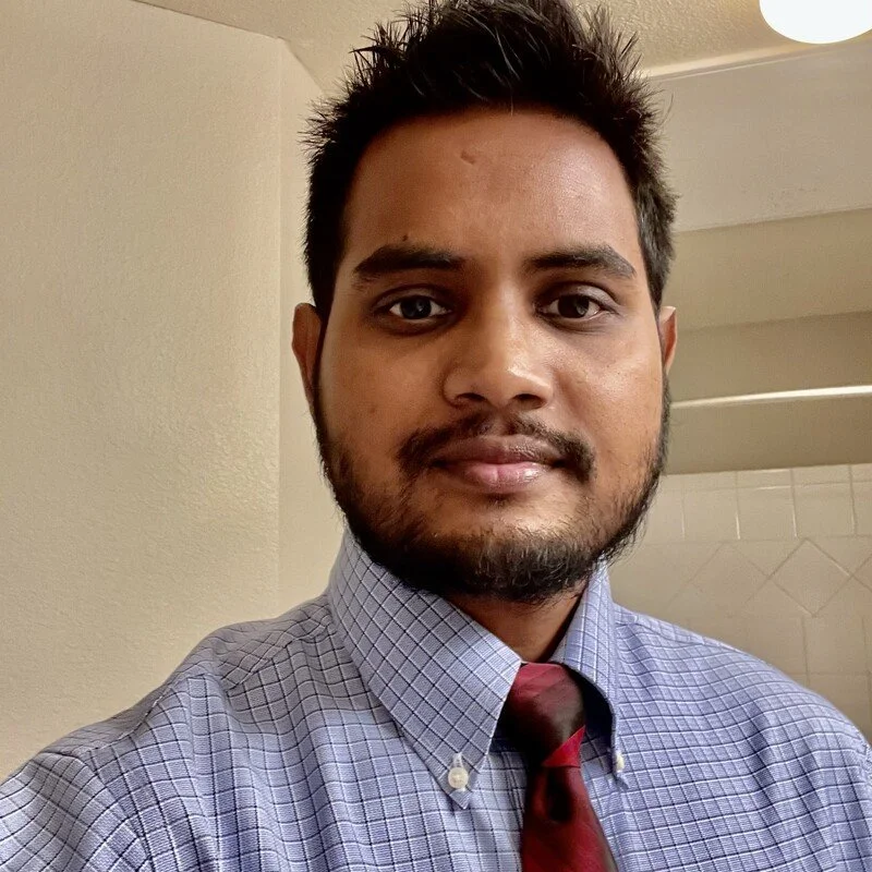 A man with short dark hair and a beard, wearing a blue checkered dress shirt and a maroon tie, standing indoors with a neutral-colored wall and ceiling with a light fixture in the background.