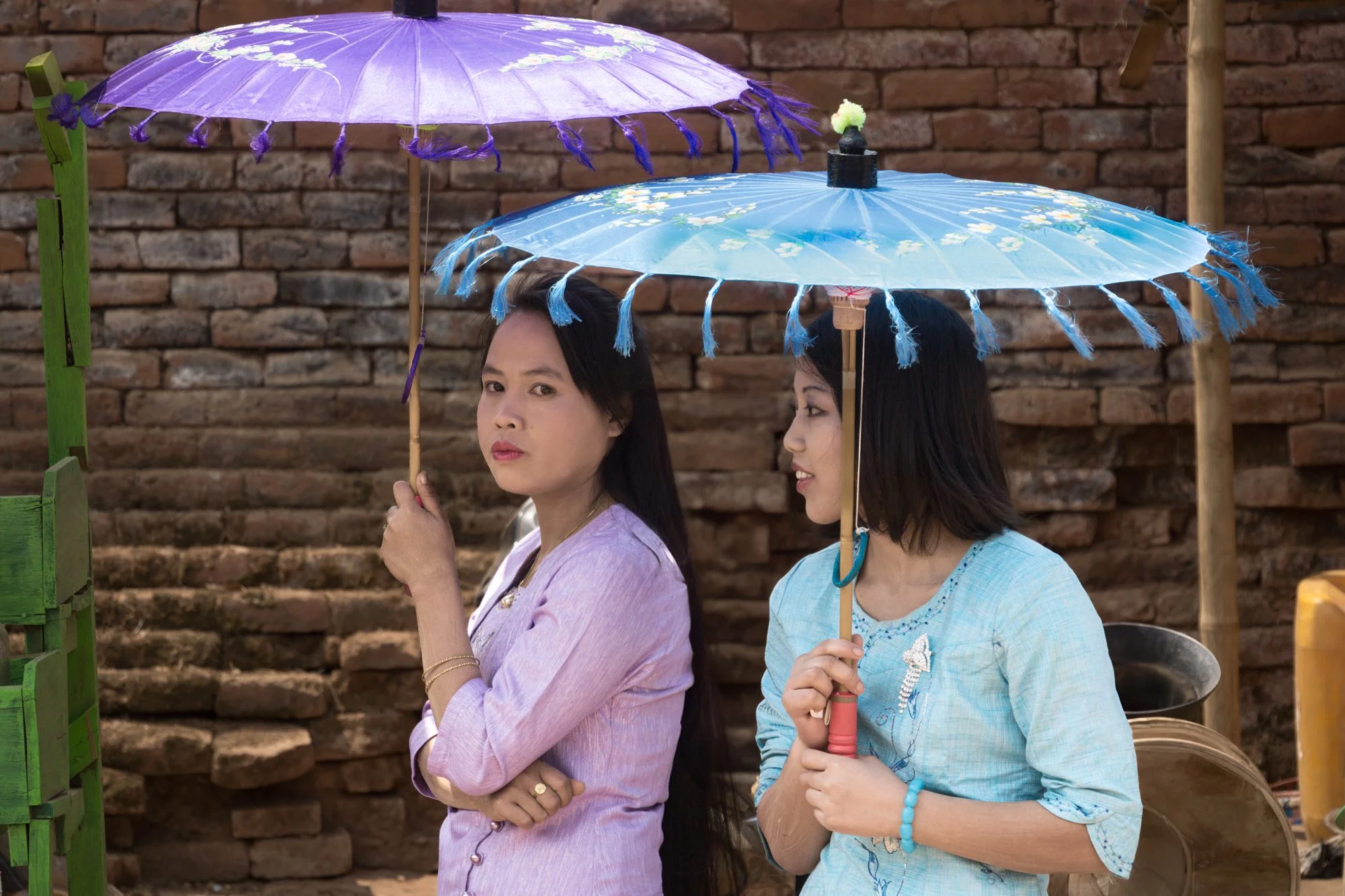 Zwei Frauen halten bunte Regenschirme, eine trägt einen lilafarbenen und die andere einen blauen, vor einem alten Backsteingebäude.