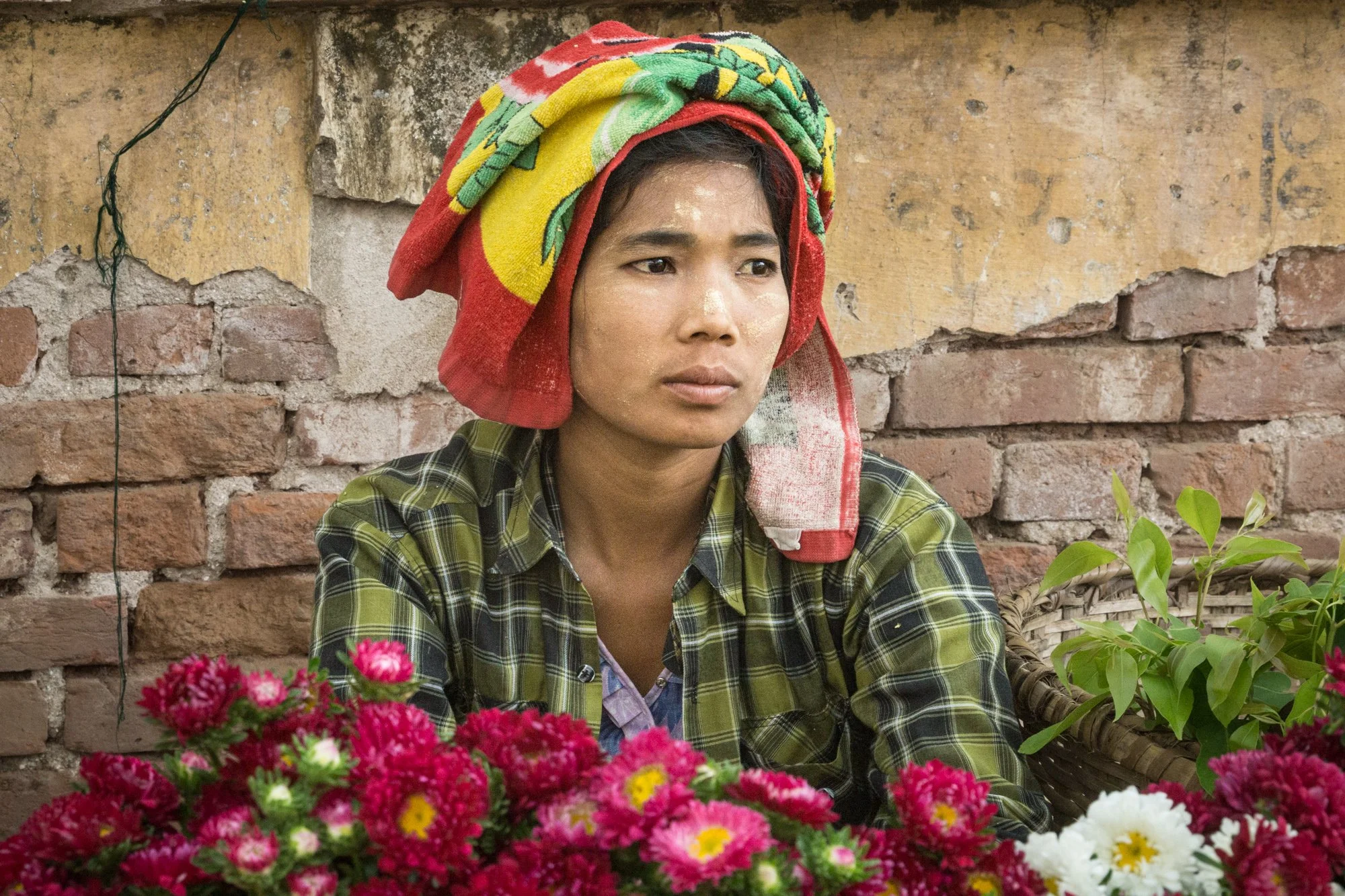 Frau mit buntem Handtuch auf dem Kopf sitzt hinter einem Blumenstand und blickt nach vorne, vor einer Backsteinmauer.