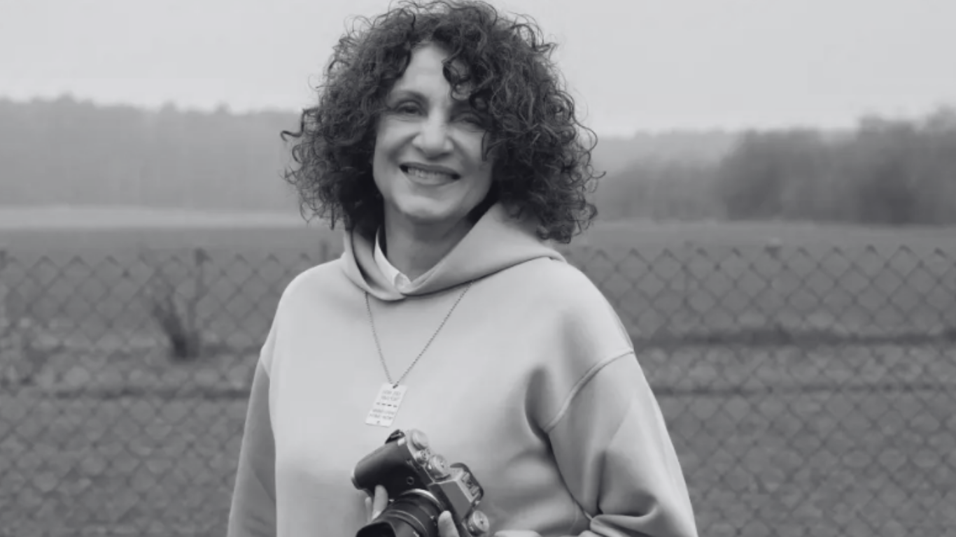 Frau mit curly Haaren, grauer Hoodie, Halsschmuck mit Schild, hält Kamera, lächelt an der Kamera, im Freien hinter einem Weidenzaun und Bäumen.