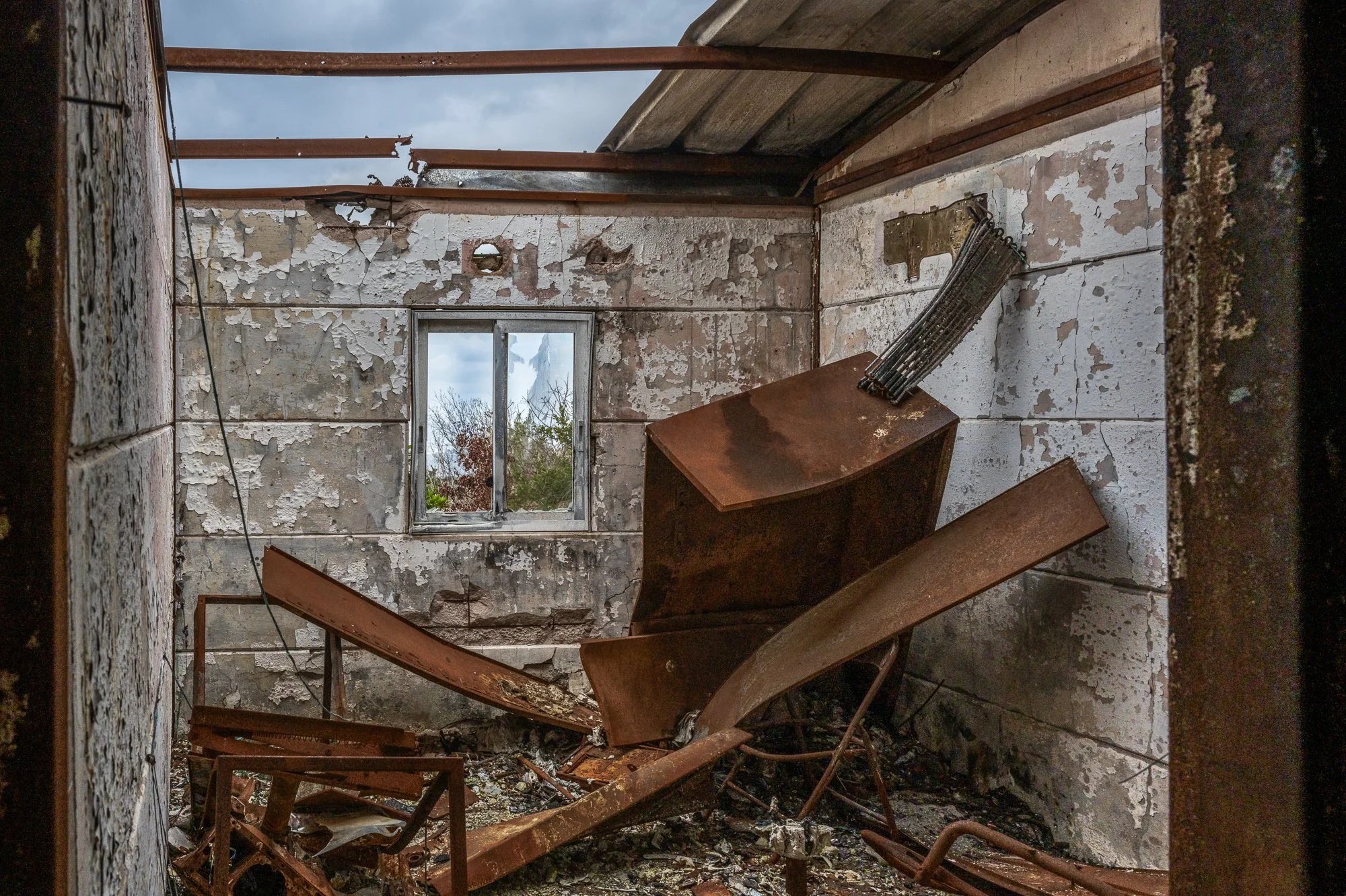 Ein verfieles, verlassenes Zimmer mit beschädigten Wänden, zerbrochenem Fenster und einem umgestürzten, verrosteten Trage. Der Himmel ist bewölkt sichtbar durch das Dachloch.