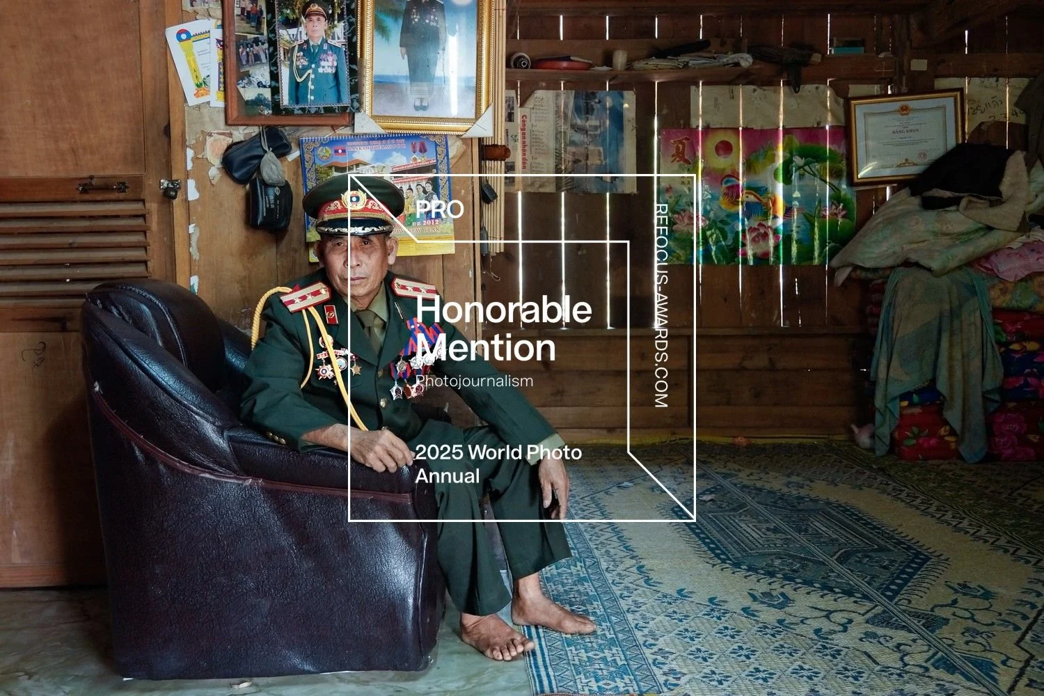 Ein älterer Mann in Militäruniform sitzend in einem rustikalen Raum mit Holzregalen, Bildern und Dekorationen an den Wänden, auf einem braunen Sessel, barfuß.