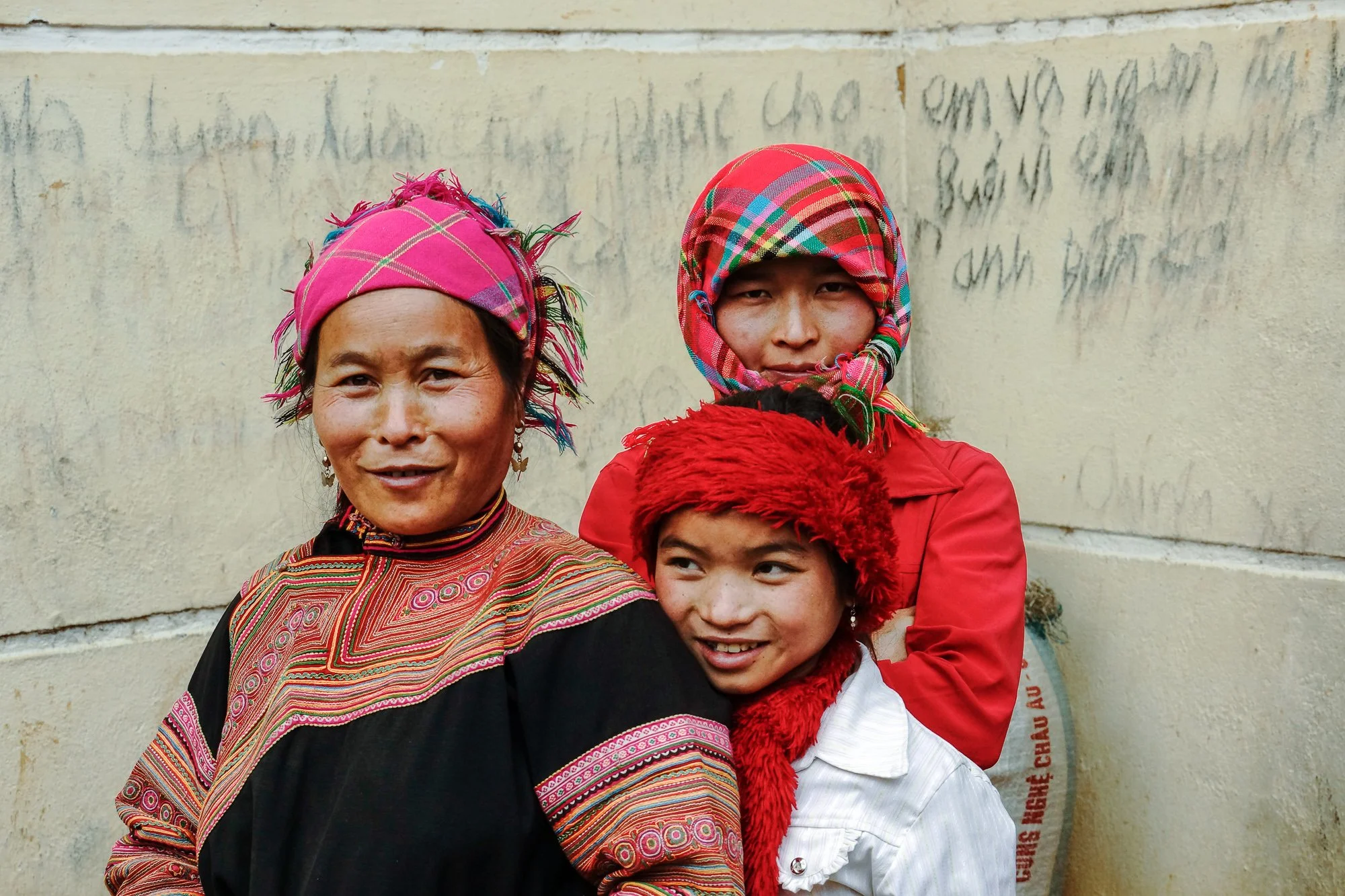 Drei Menschen in traditioneller Kleidung und bunten Kopfbedeckungen, vor einer alten Wand mit Graffiti.