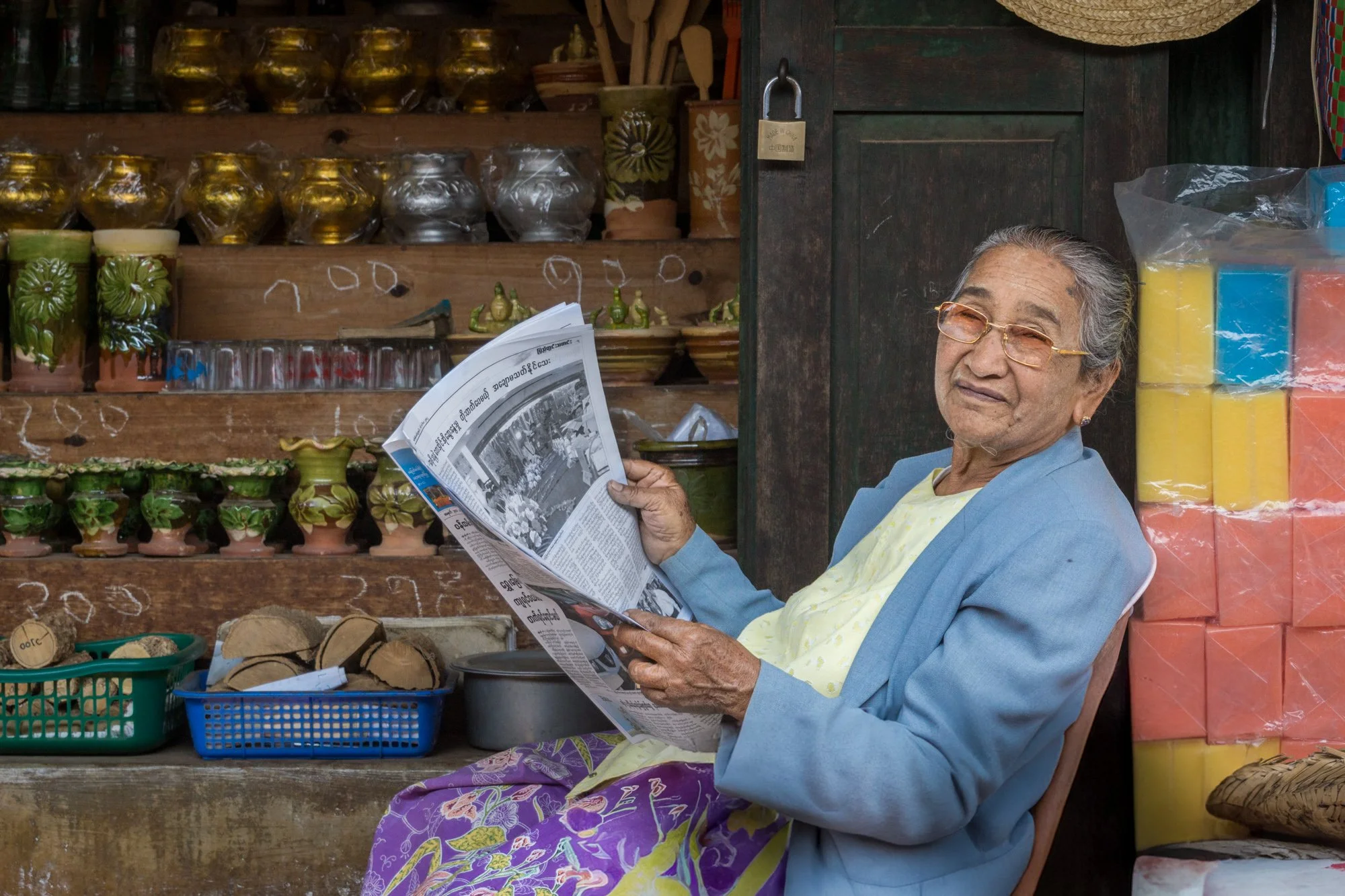 Ältere Frau mit Brille liest Zeitung in einem Laden, der handgefertigte Töpferwaren und bunte Schwämme verkauft.