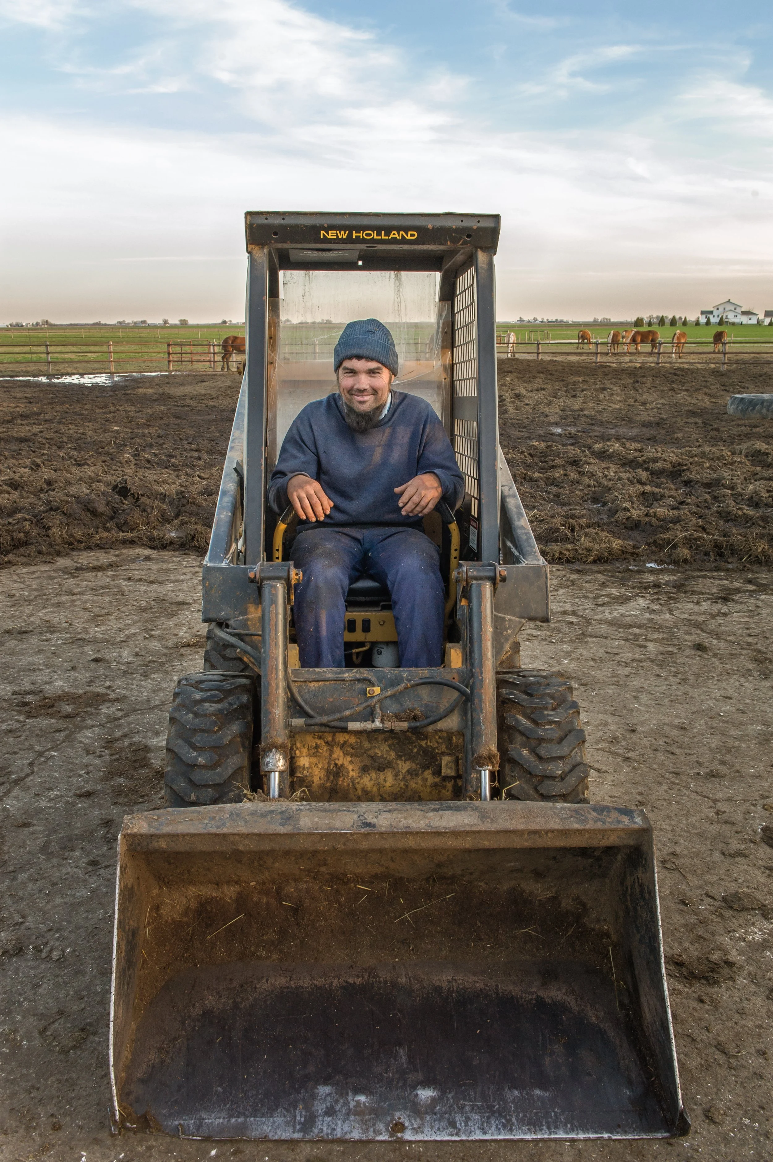 Mann sitzt auf einem kleinen Traktor auf einem Bauernhof, im Hintergrund Weide mit Pferden und ein Haus unter einem bewölkten Himmel.