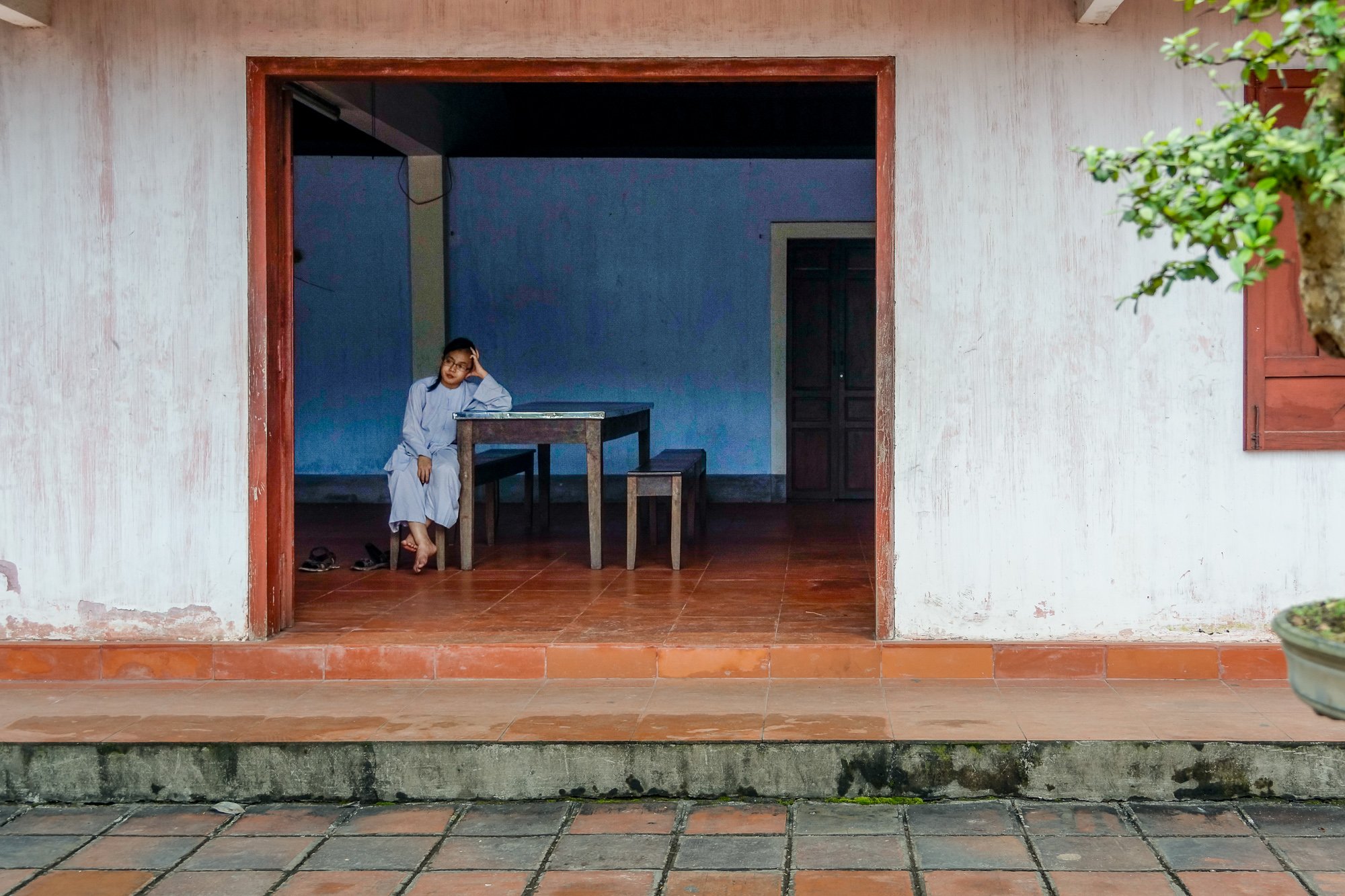 Ein junges Mädchen in weißer Kleidung sitzt an einem Holztisch in einem offenen Raum, der Teil eines Hauses ist. Der Innenraum hat blaue Wände und einen dunklen Türrahmen, auf dem Boden liegen Schuhe. Draußen vor dem Raum ist eine Terrasse mit roten Ziegeln und Pflastersteinen, und ein Baum ist sichtbar auf der rechten Seite.