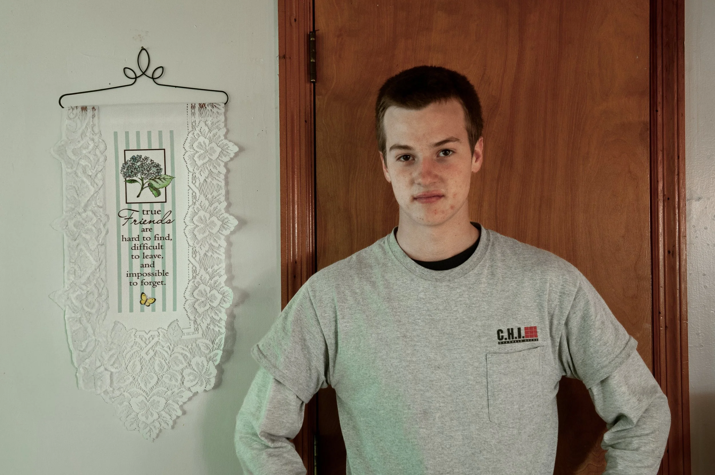 Ein junger Mann mit kurzen braunen Haaren trägt ein graues Sweatshirt vor einem Holztür. An der weißen Wand links hängt ein handgeschnitztes, weißes Läuferbordüre-Teppich mit einer gedruckten Nachricht und einem Bild eines Baums, Blatt und Schmetterlings.
