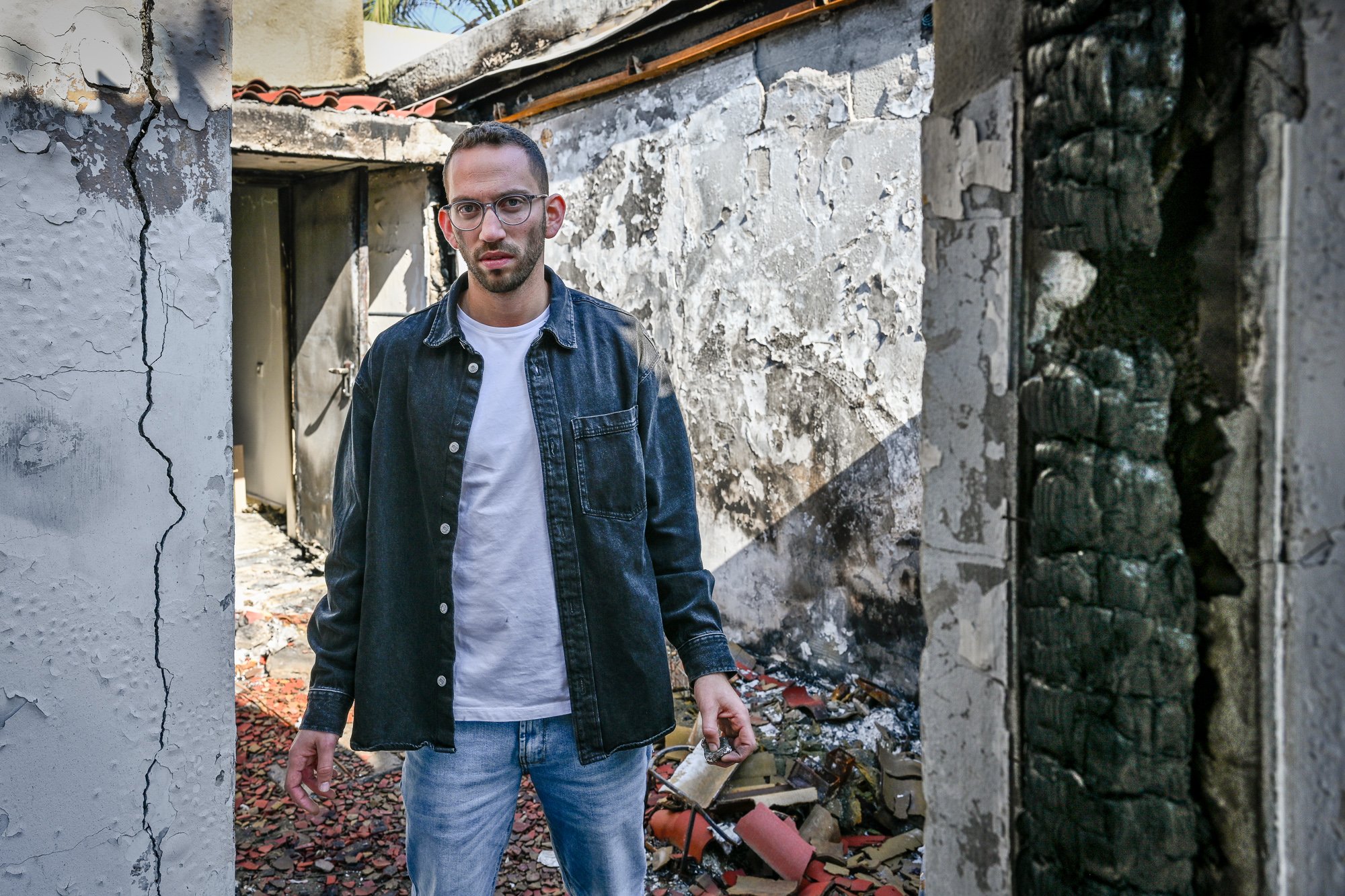 Junger Mann mit Brille, Jeansjacke und weißem T-Shirt steht in einer zerstörten, brennenden Umgebung, umgeben von Brandschäden und Trümmern.