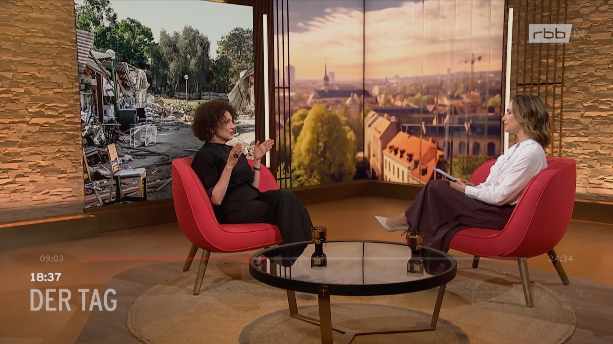 Zwei Frauen sitzen in einem Studio-Interview, im Hintergrund ist eine Kulisse mit einer Stadtansicht bei Sonnenuntergang und nebenan eine zerstörte Nachbarschaft zu sehen.