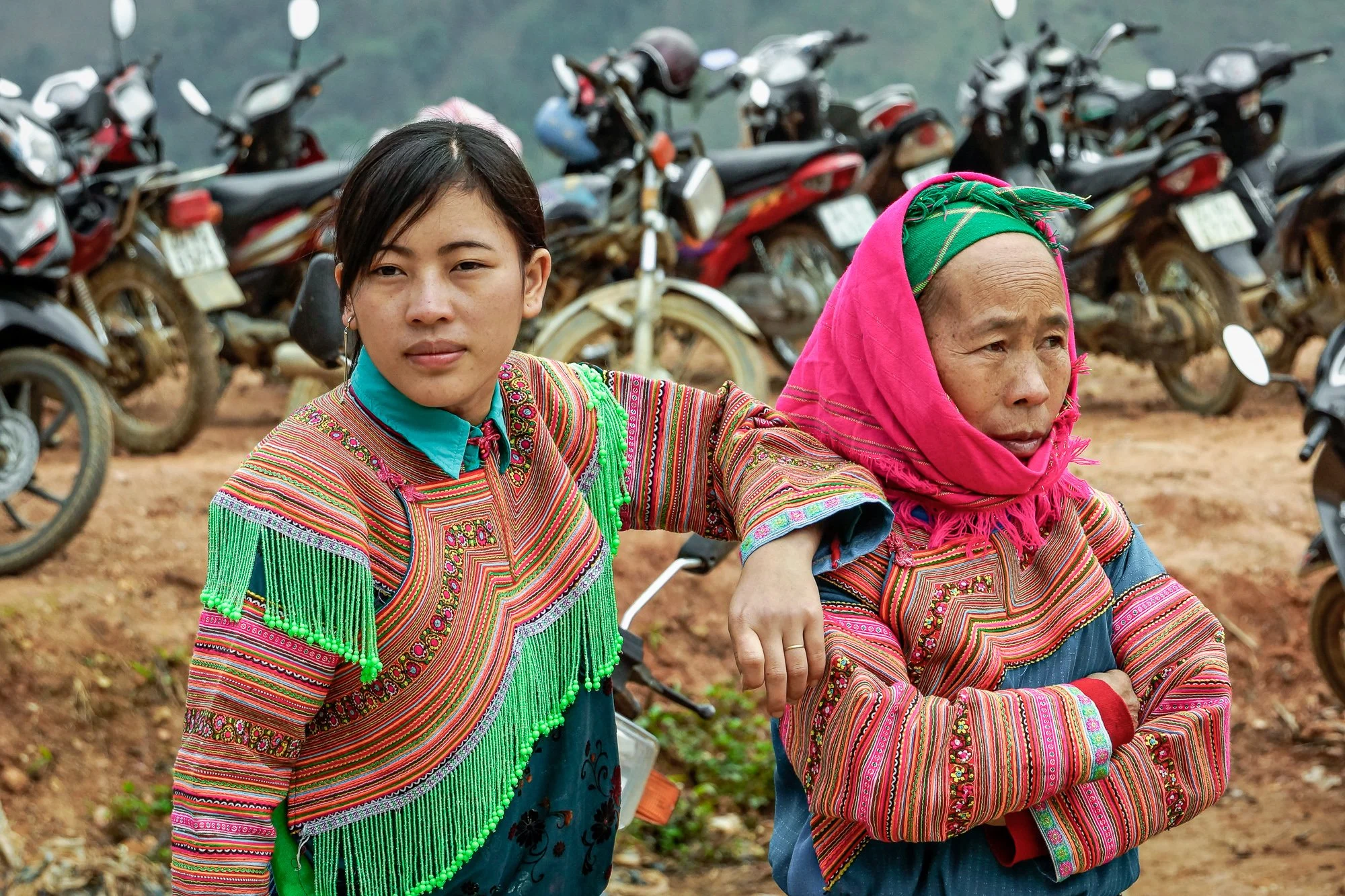 Zwei Frauen in traditionellen, bunten Trachten mit Schnitten und Stickereien, vor einem Hintergrund aus Motorrädern auf unbefestigtem Boden.