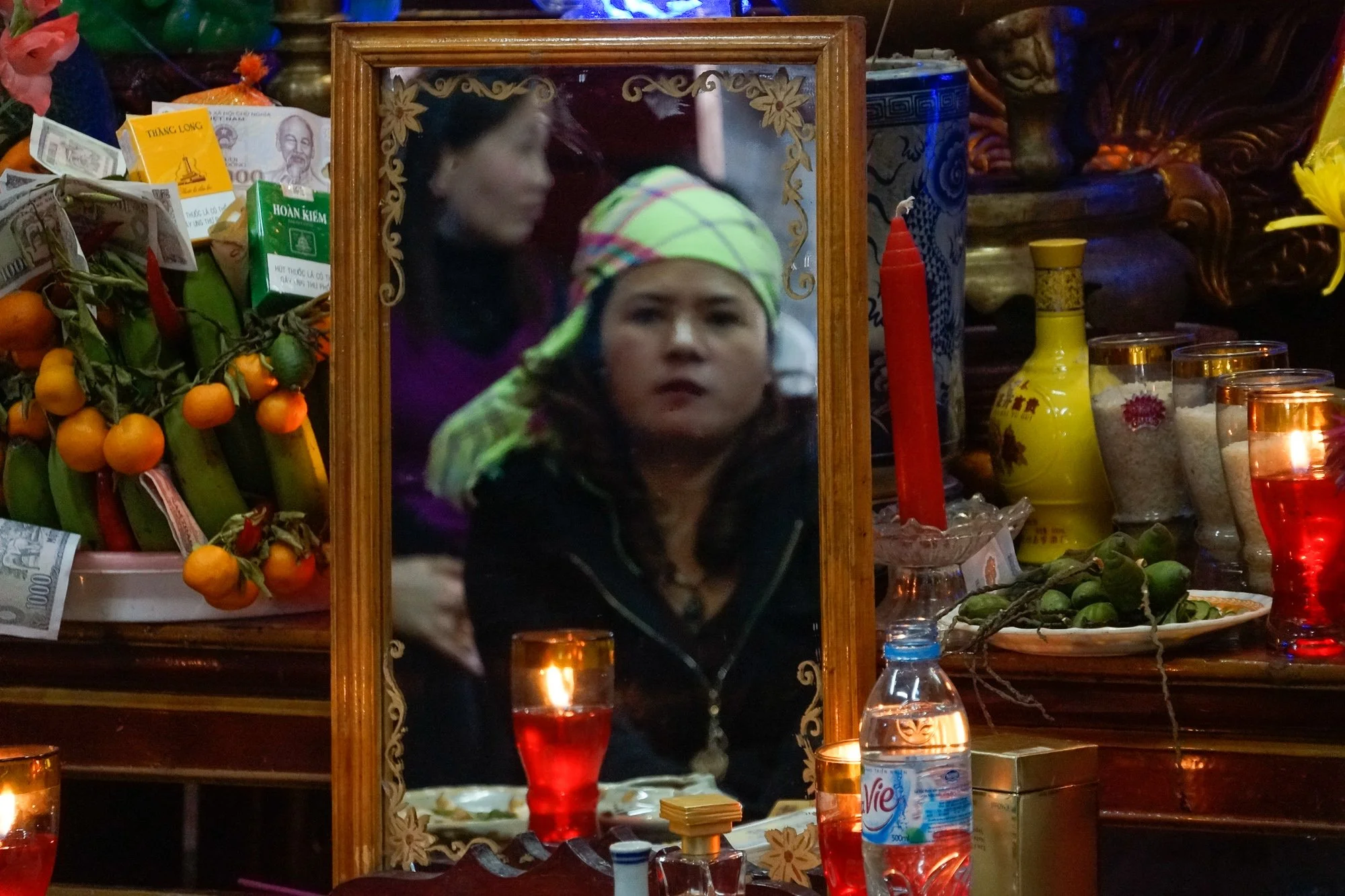 Ein Spiegel mit Holzrahmen, der eine junge Frau mit dunklen Haaren und einem bunten Schal auf dem Kopf zeigt. Im Hintergrund sind Erinnerungsstücke, Kerzen, Obst und Getränke auf einem altarartigen Tisch zu sehen.