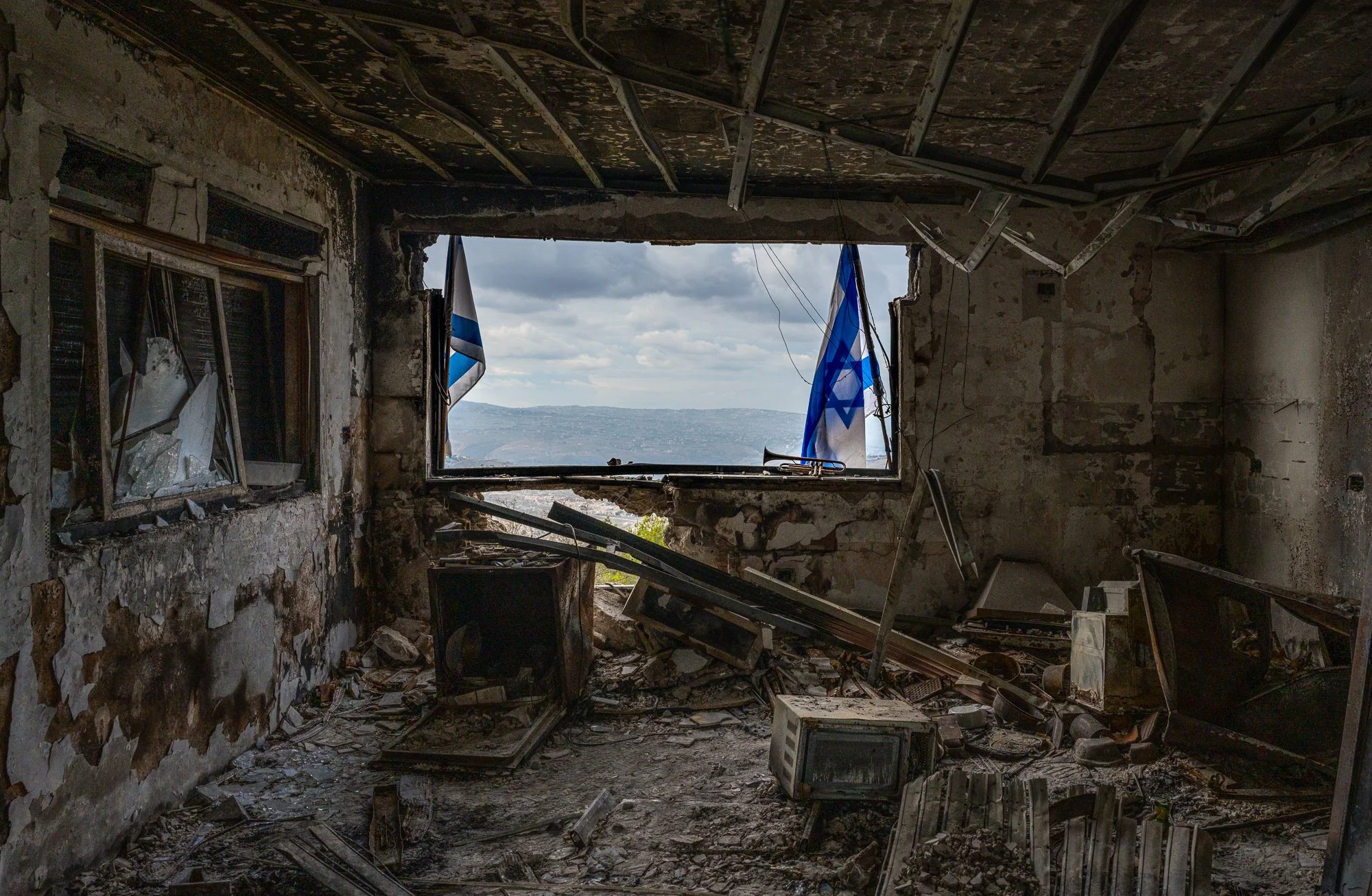Ein zerstörter Raum mit einem großen, offenen Fenster, durch das man den Himmel und Berge sieht, sowie zwei israelische Flaggen. Der Raum ist verwüstet mit zerbrochenen Fernsehern, Trümmern und beschädigter Wanddekoration.