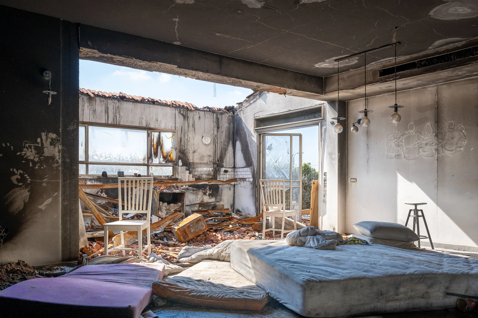 Innenraum eines zerstörten, brandschattigen Hauses mit zerbrochenen Fenstern, einem beschädigten Bett, zerlegten Möbeln und Schutt auf dem Boden.