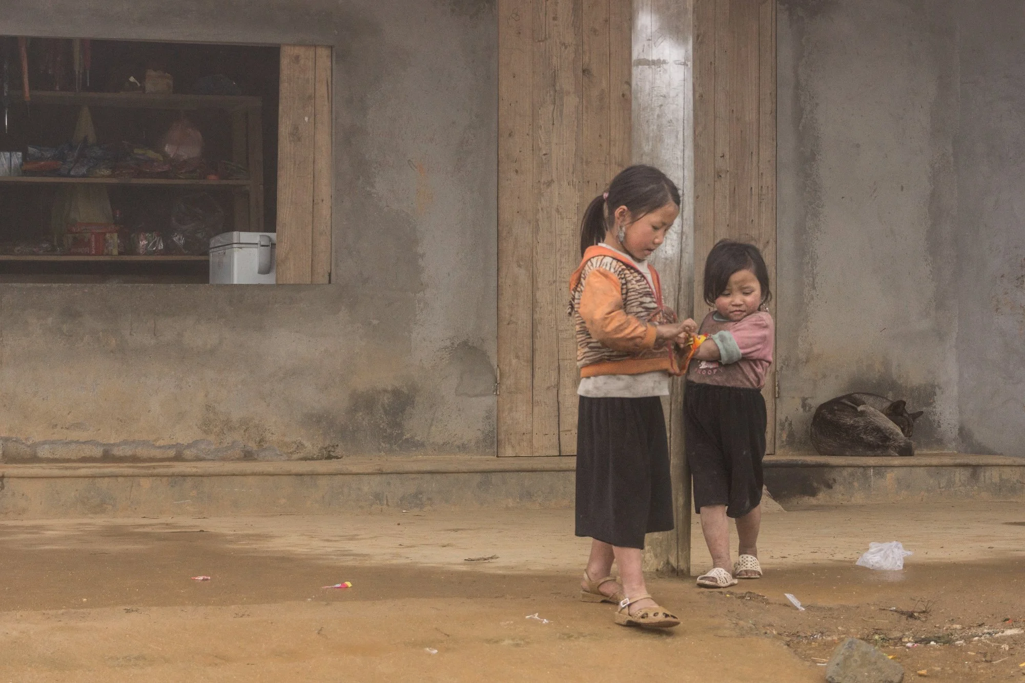 Zwei junge Mädchen stehen in einem schlichten, ungepflegten Raum mit rohem Bodengrund und einer Wand aus Beton und Holz. Eines der Mädchen hält ein Objekt in den Händen, während das andere sie ansieht. Im Hintergrund liegt eine Katze auf dem Boden, seitlich befindet sich eine ungeordnete offene Holzregal mit Gegenständen.
