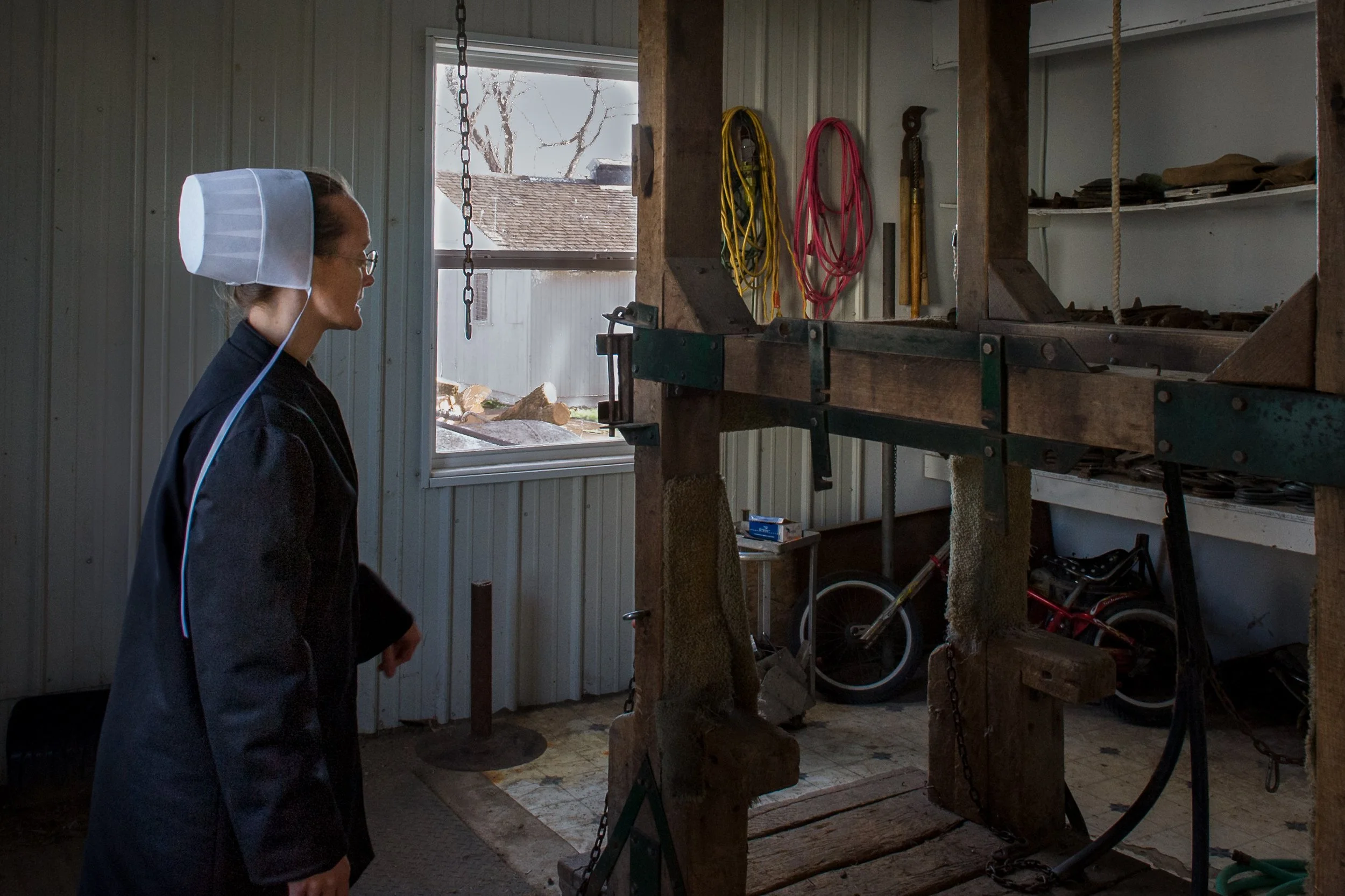 Eine Frau in schwarzem Kleid und Haube steht in einer Werkstatt, schaut auf ein großes handwerkliches Holz- oder Metallgestell. Die Werkstatt hat eine weiße Wandpaneele, ein Fenster, das Tageslicht einlässt, und Werkzeuge an der Wand hängen.