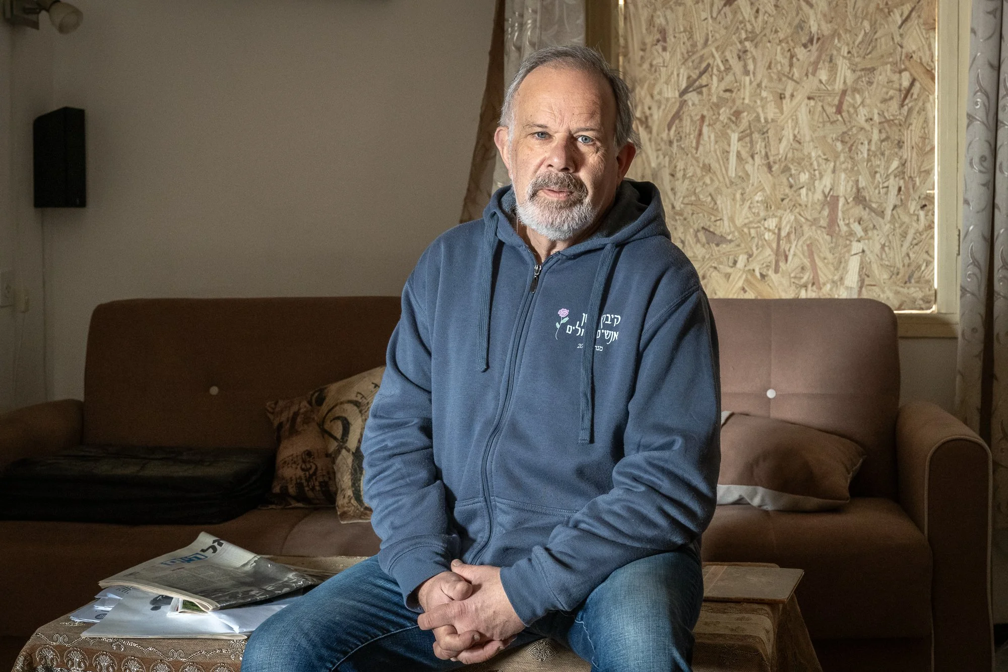 Ein älterer Mann mit grauem Bart sitzt auf einem Sofa in einem ungefederten Wohnzimmer, trägt eine dunkle Kapuzenjacke und schaut direkt in die Kamera.