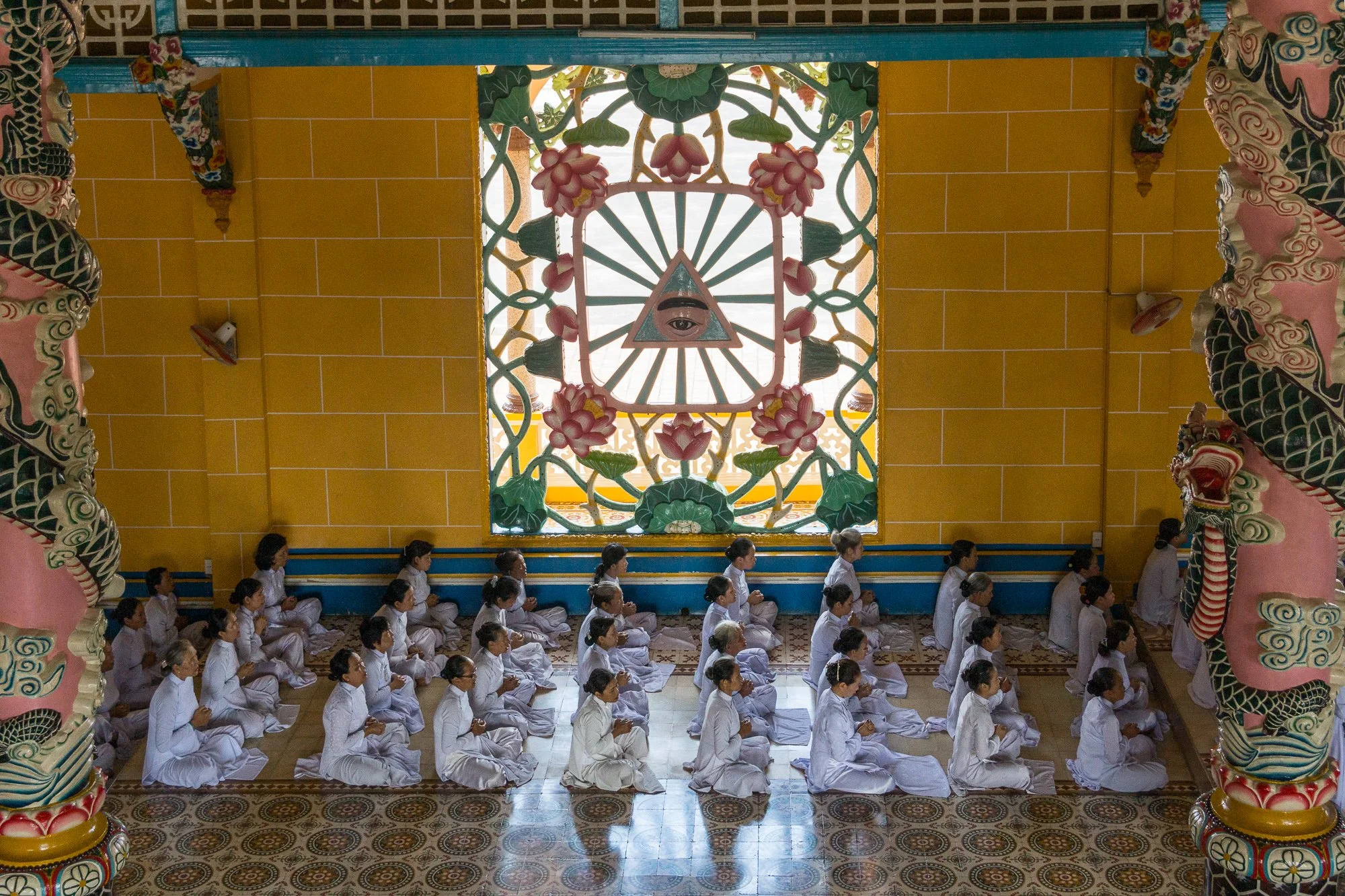 Menschen, die in einem Tempel in weißen Kleidung sitzen und meditieren, im Hintergrund ein großes farbenfrohes Fenster mit einem Auge in einem Dreieck, umgeben von Lotusblumen.