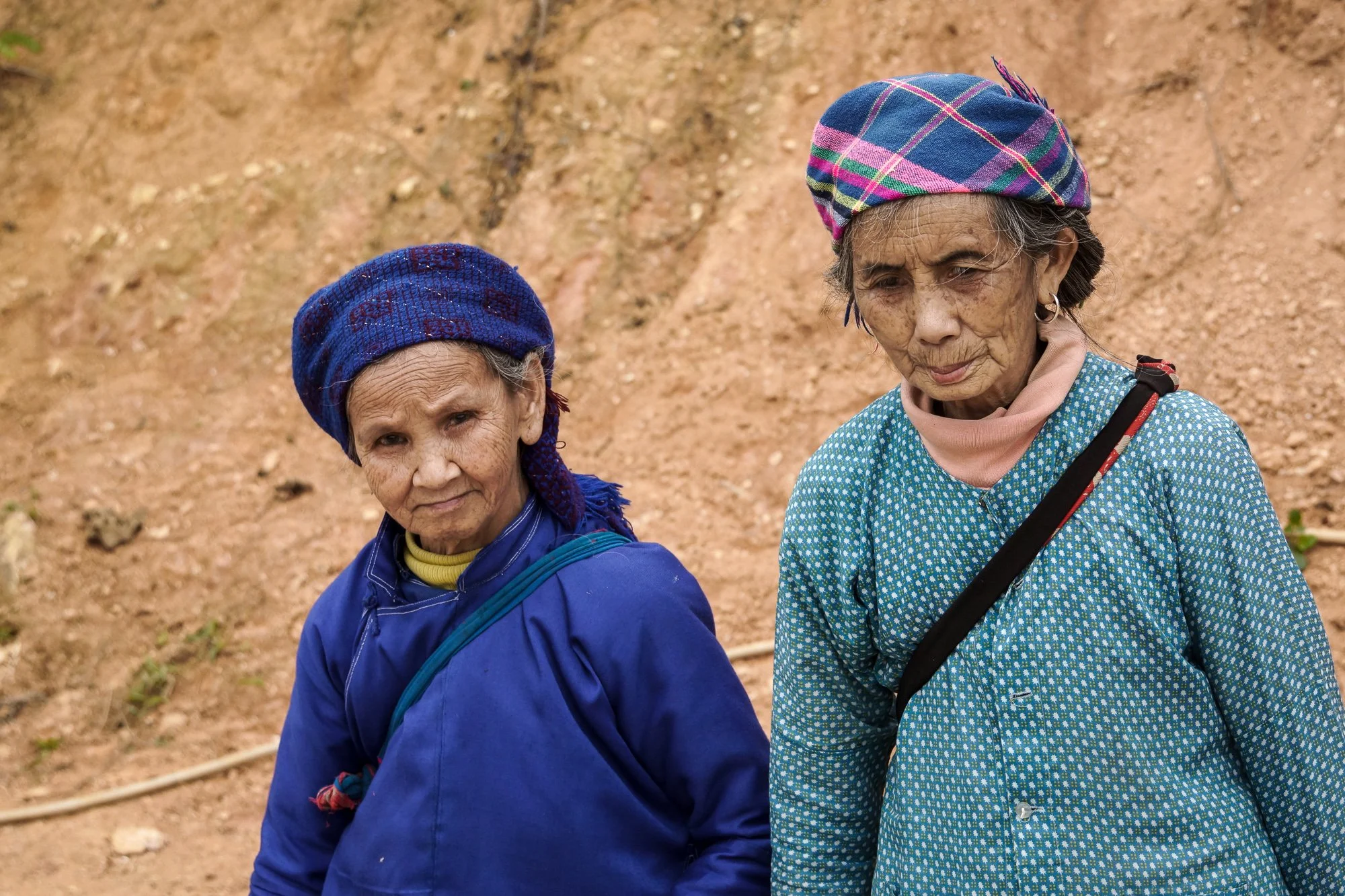 Zwei ältere Frauen mit traditioneller Kleidung und bunten Tüchern auf dem Kopf stehen vor einem erdigen Hintergrund.