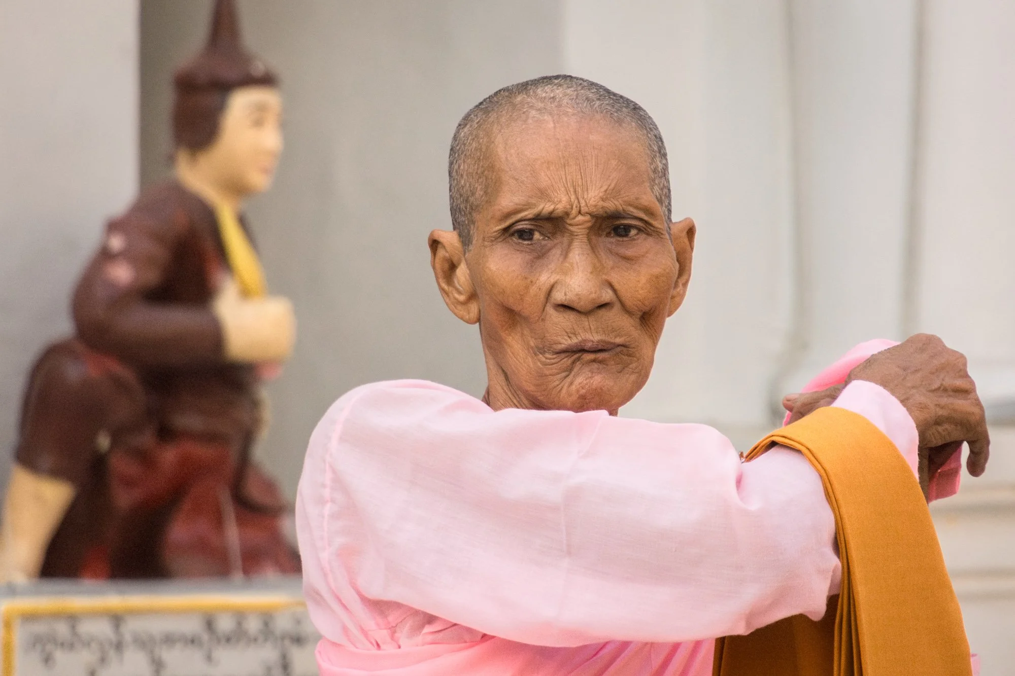 Ein älterer Mann in traditioneller Kleidung schaut ernst. Im Hintergrund eine Buddha-Statue.
