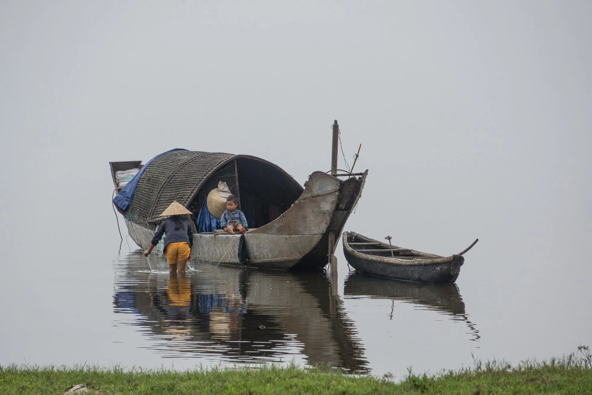 Zwei Boote liegen im Wasser, eine Person steht im Wasser und wäscht sich, während ein Kind im größeren Boot sitzt. Das kleinere Boot ist neben dem großen Boot verankert.