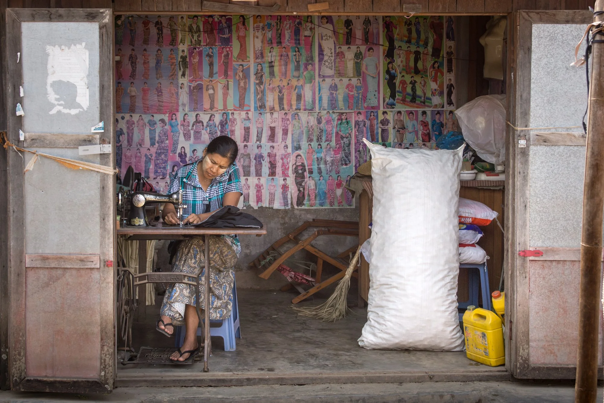 Eine Frau arbeitet an einer Nähmaschine in einem kleinen, einfachen Raum, der mit bunten Postern mit Modedarstellungen an der Wand dekoriert ist, im Hintergrund befinden sich verschiedene Gegenstände, darunter große Säcke, Reinigungsbürsten und Reinigungsmittel.