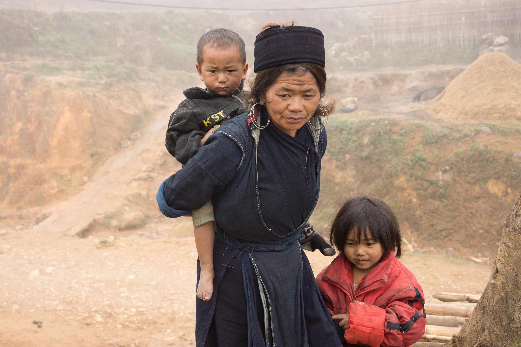 Frau mit einem Kind auf dem Rücken und einem weiteren Kind neben ihr in einer staubigen, ländlichen Umgebung.