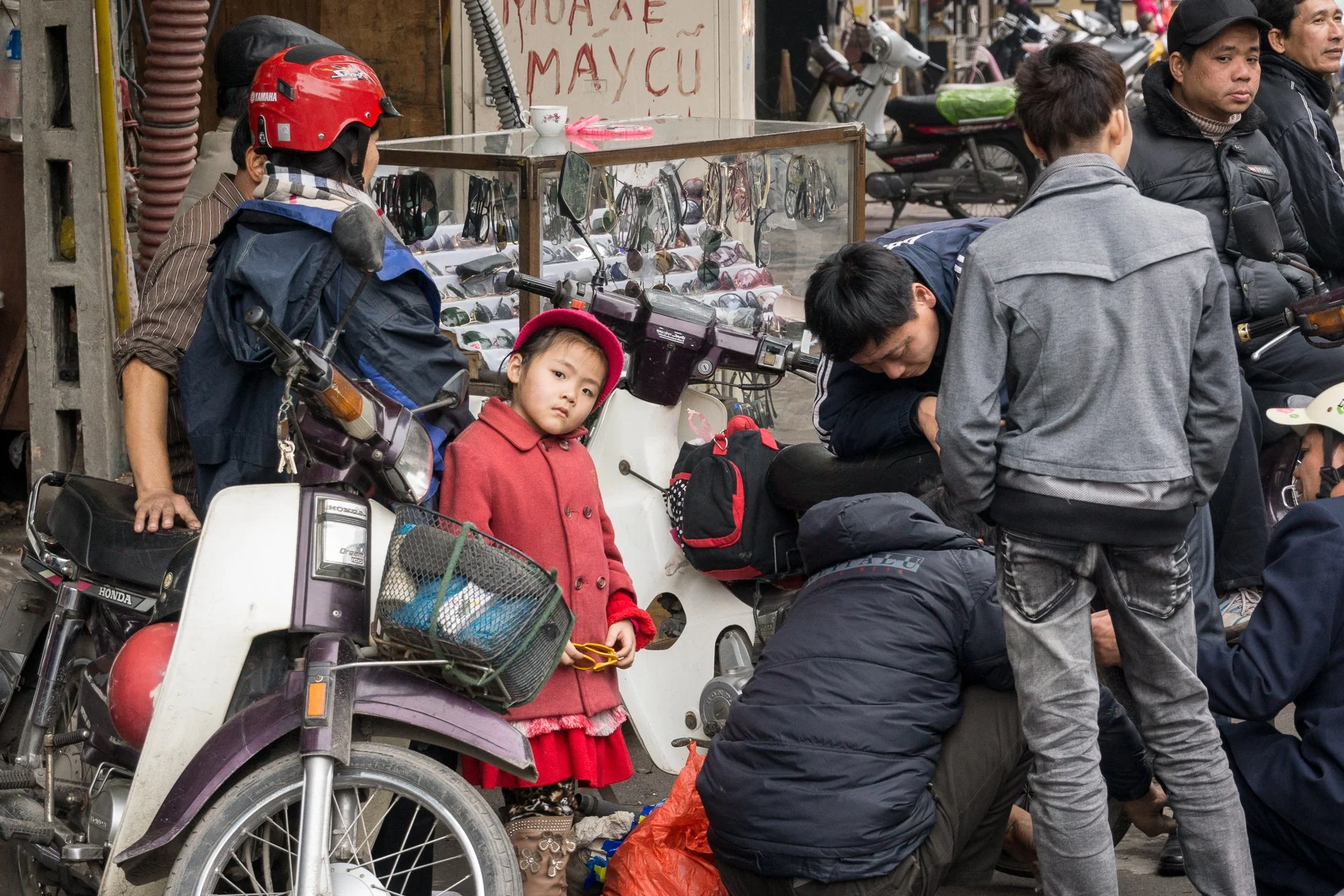Junge in rotem Mantel und roter Hut steht neben Motorrädern und einem Straßenstand, umgeben von Menschen, einige im Gespräch oder bei Reparaturen.