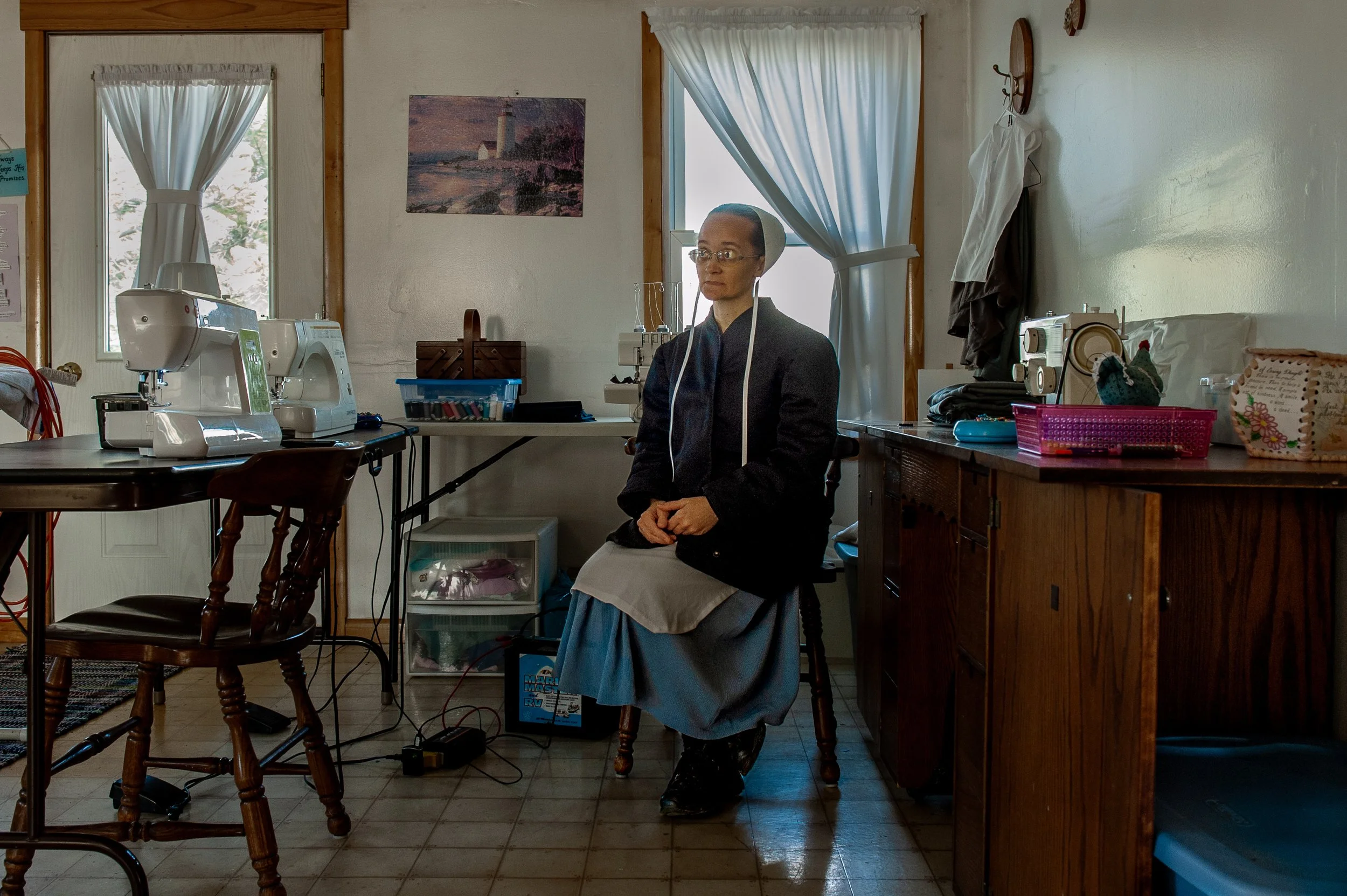 Eine Frau in alter Kleidung sitzt in einem Raum mit verschiedenen Nähmaschinen und anderen Textilwerkzeugen, die auf Tischen stehen. Sie trägt eine Haube und scheint an einer Nähmaschine zu arbeiten oder zu warten.