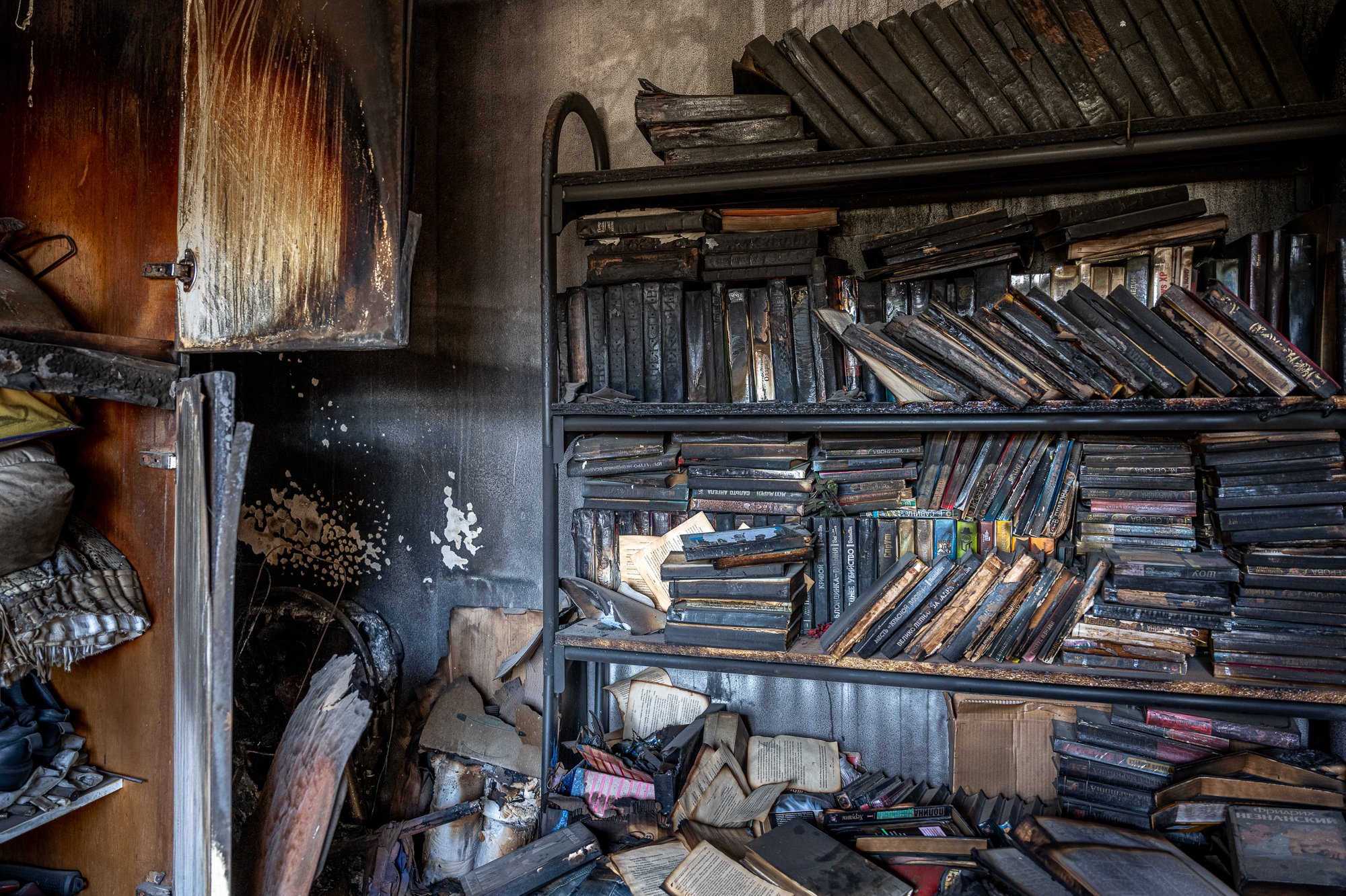 Bücherregal mit verbrannten, verkohlten Büchern in einem Raum, daneben zerbrochene Kartons und verbrannte Gegenstände.