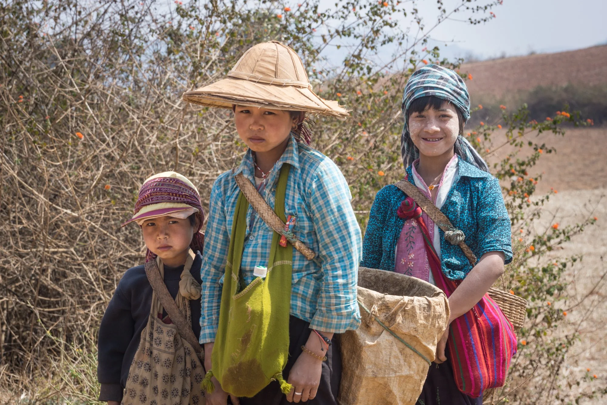 Drei junge Mädchen mit traditioneller Kleidung und Sonnenhüten in einer ländlichen Umgebung, umgeben von Büschen und niedrigen Hügeln.